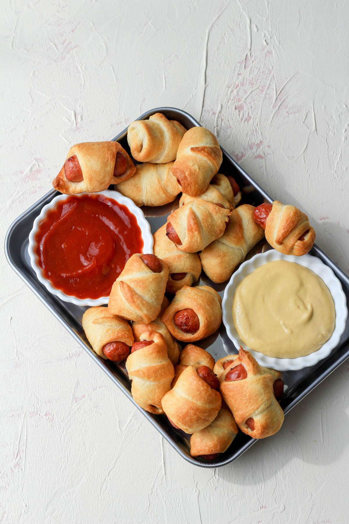 A small silver tray on a cream counter with air fryer pigs in a blanket and a bowl of ketchup in the left and mustard in the right.