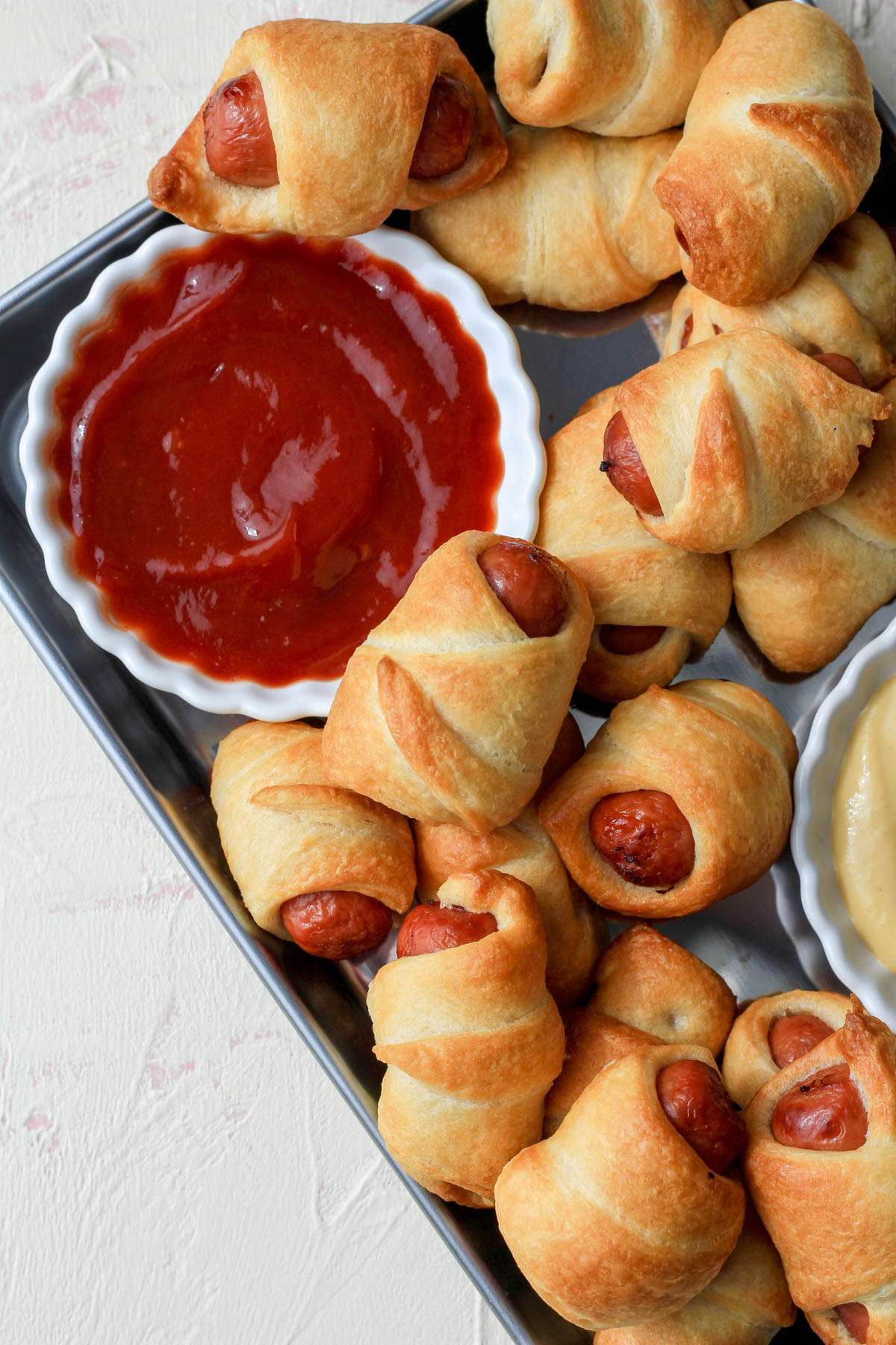 A corner of a silver pan filled with air fryer pigs in a blanket surrounding a white bowl of ketchup.