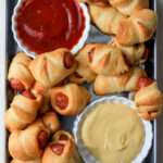 A close up of a silver pan with pigs in a blanket and a bowl of ketchup in the top left and mustard in the bottom right.
