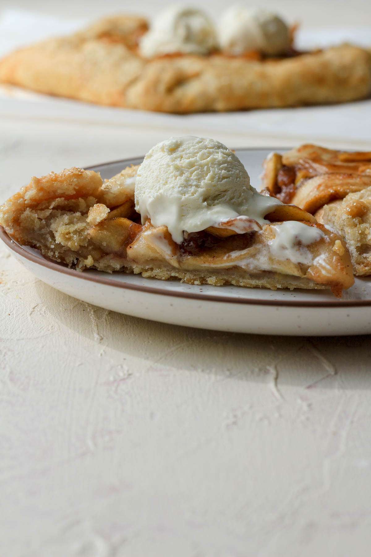 A white plate with a brown rim with a slice of apple galette topped with a scoop of vanilla ice cream from the side.