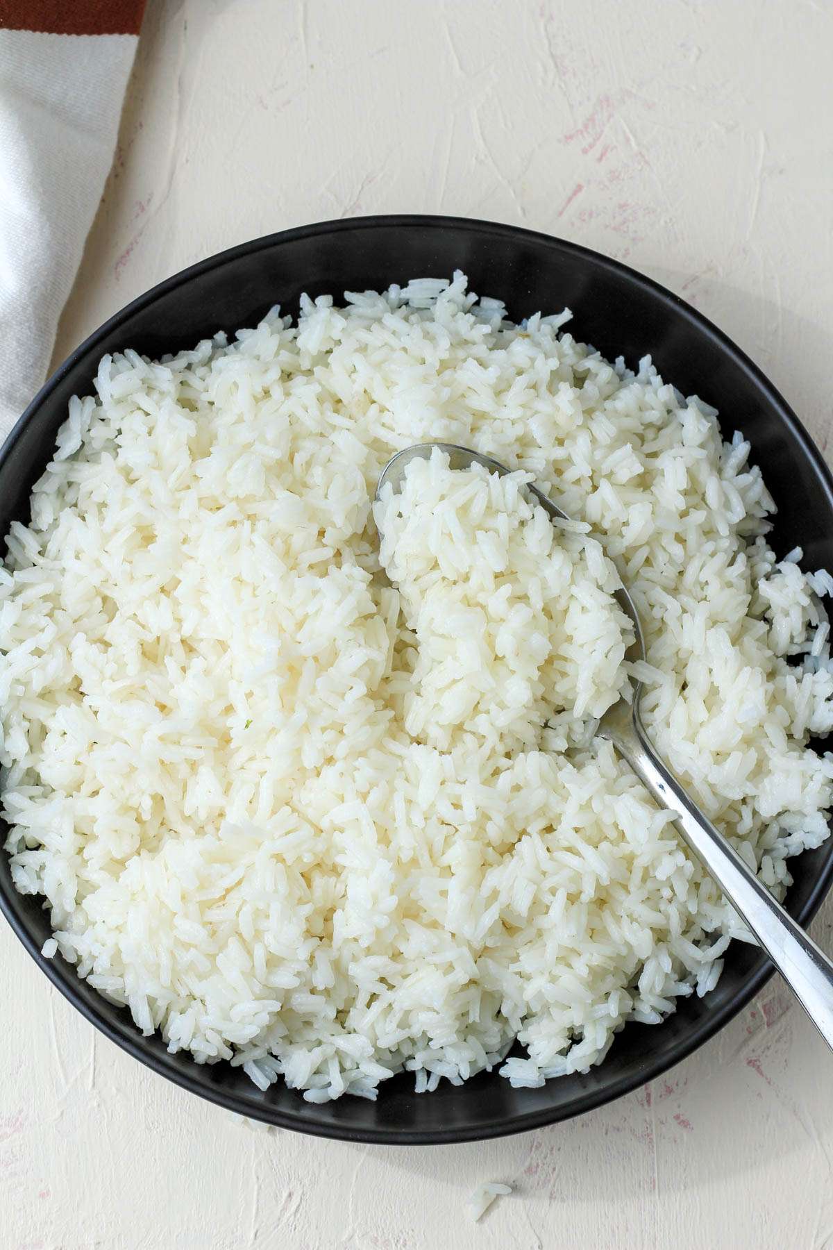 A top down picture of a black bowl with a silver spoon filled with cooked long grain rice.