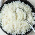 A close up of a black bowl of long grain rice with a sliver spoon scooping the rice.