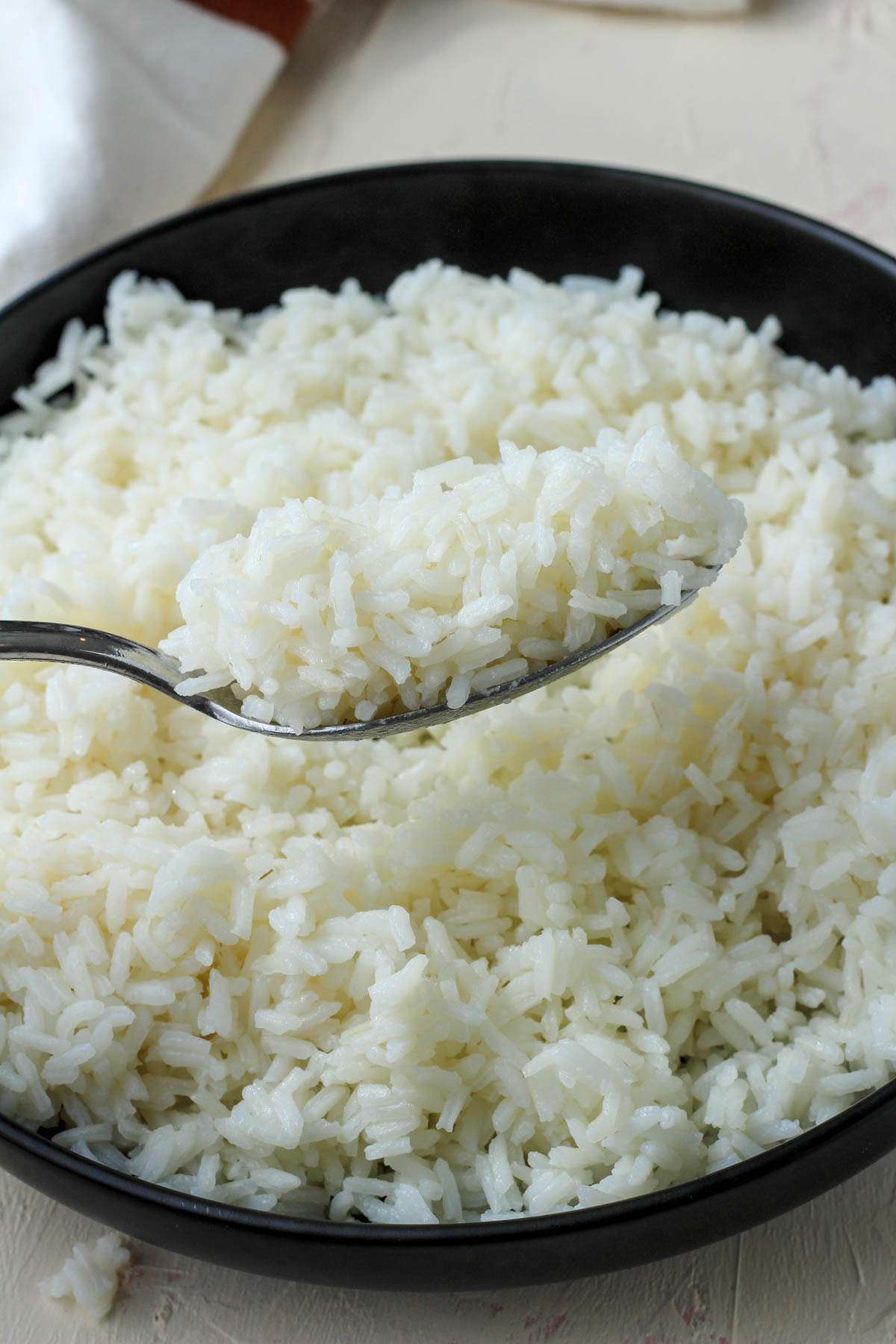 A silver spoon of long grain rice over a black bowl with cooked long grain rice.
