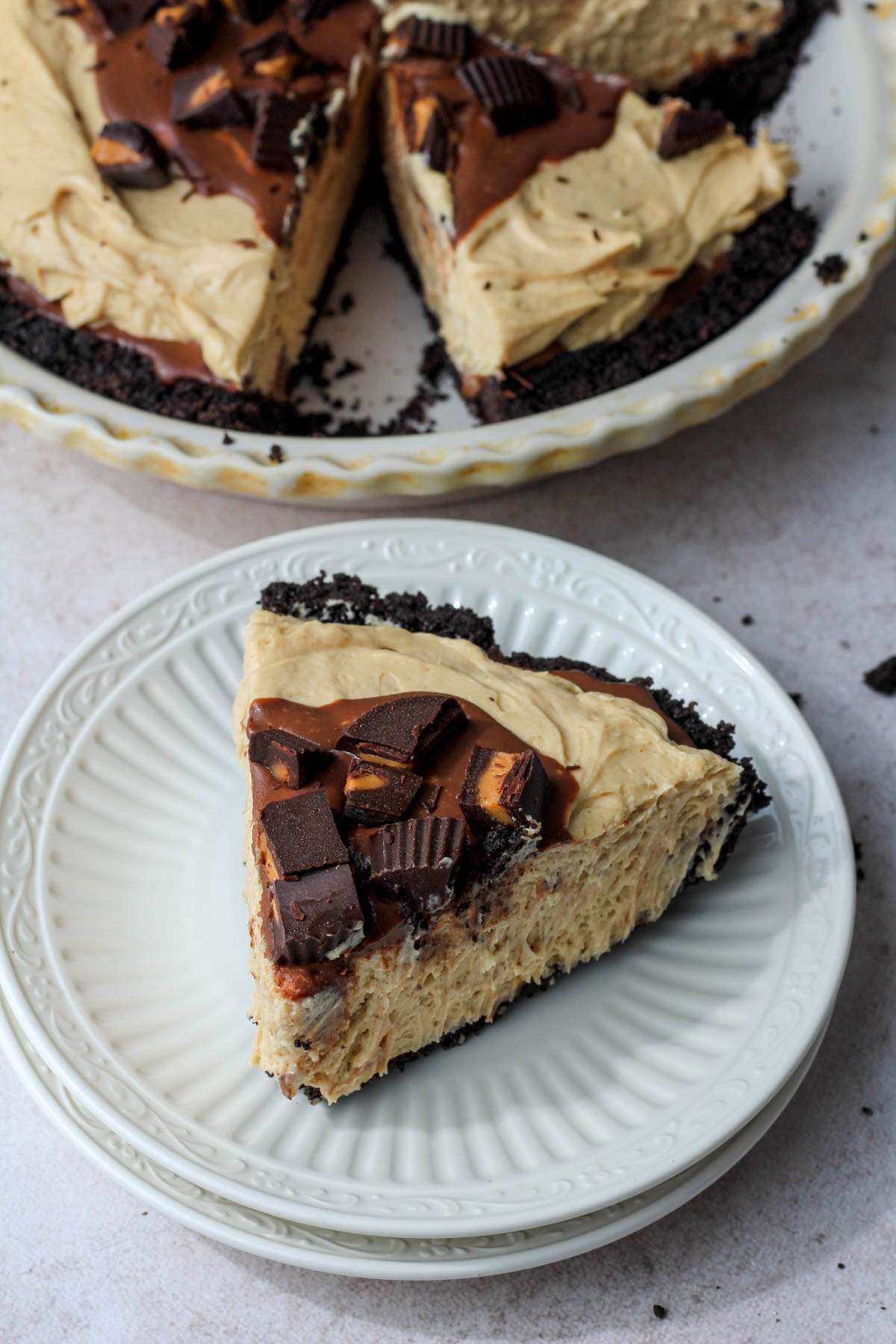 A white plate with a slice of vegan no bake peanut butter pie topped with peanut butter cup pieces in front of the pie pan.