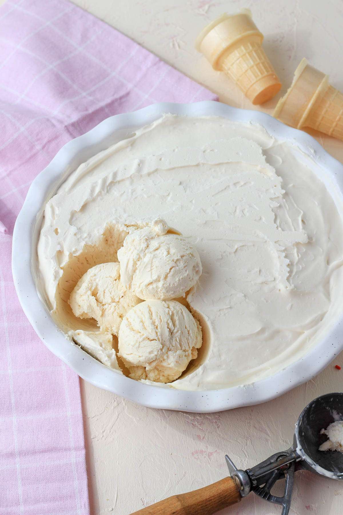 A deep dish of no churn vegan vanilla ice cream with three scoops on the left side of the pan, a few ice cream cones above the dish, and an ice cream scoop in the bottom right.