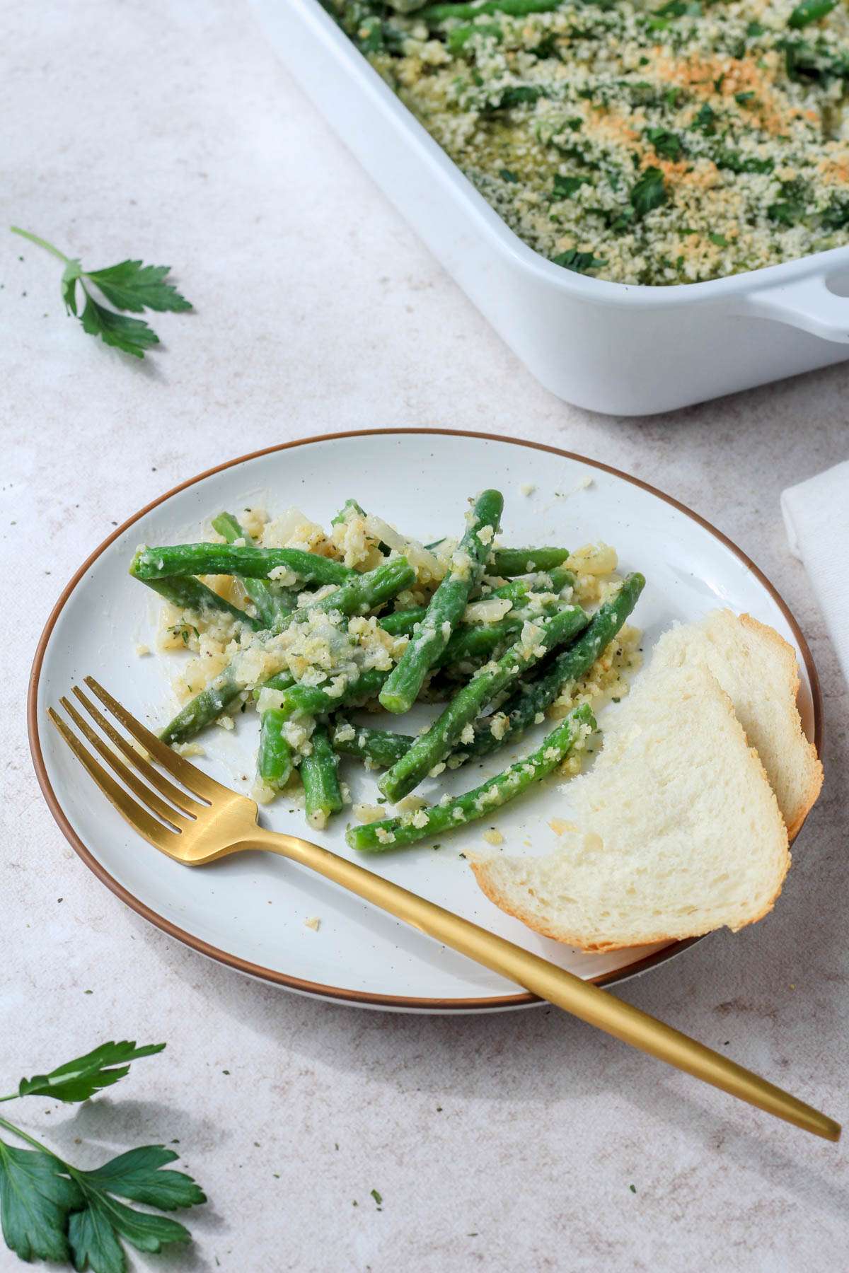 A white plate with a small serving of vegan green bean casserole with bread and a fork on the plate and the casserole dish in the back left of the photo.