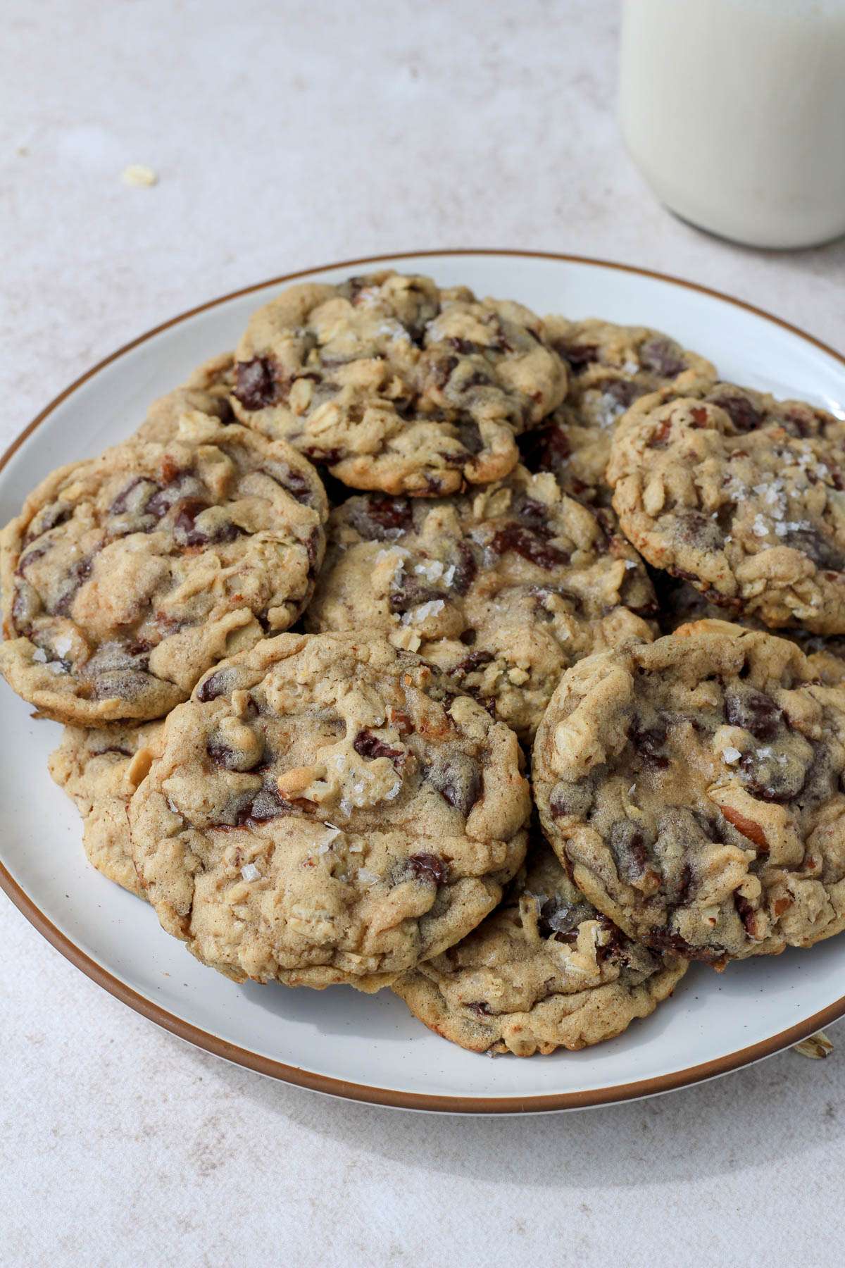 A white plate with a brown rim topped with a layer of dairy-free cowboy cookies and a small glass jug of dairy-free milk in the back right corner.