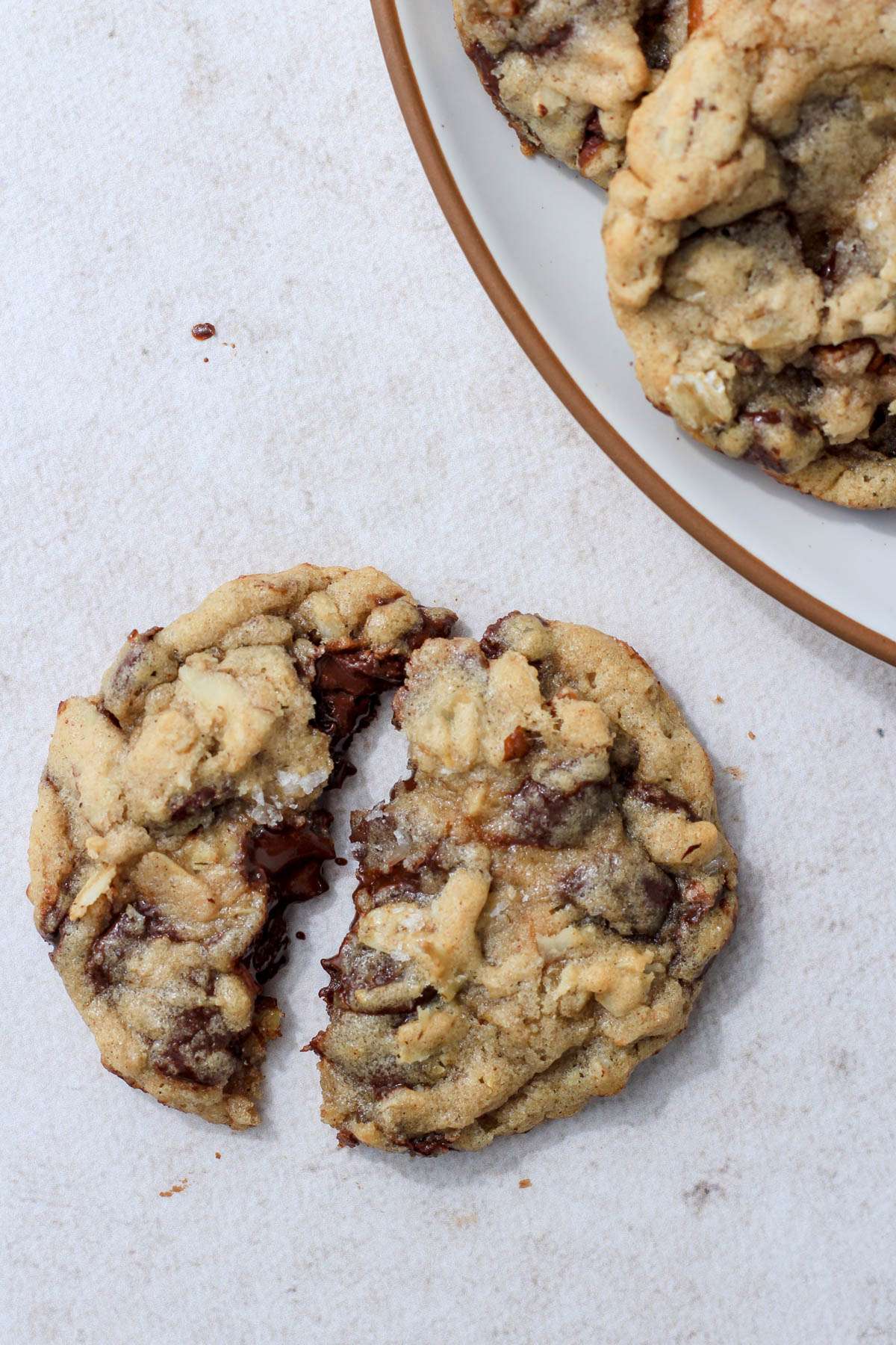 A warm cowboy cookie broken in half on a a counter next to a white plate with a brown rim topped with cowboy cookies.