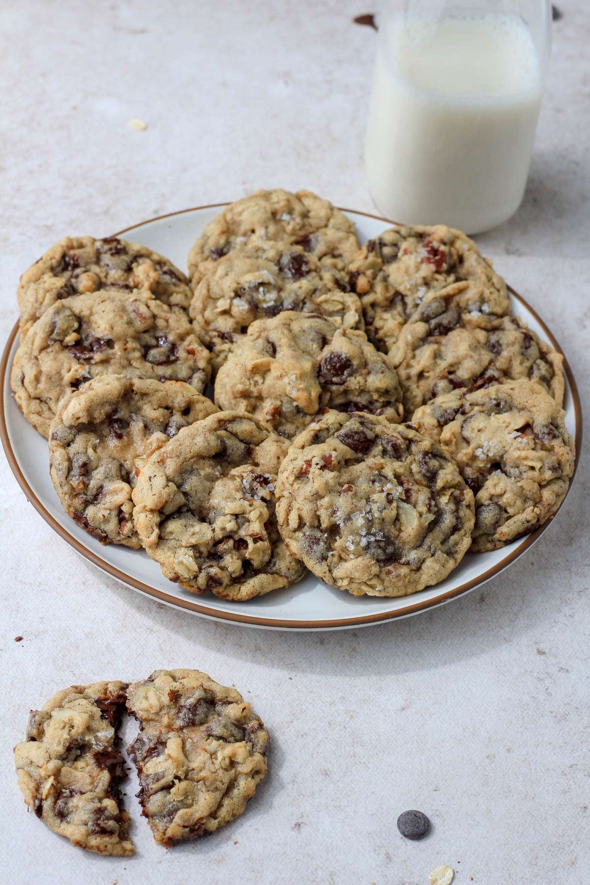 A glass jug of dairy-free milk in the top right, a white plate with a brown rim topped with cowboy cookies in the middle, and a single cowboy cookies pulled in half in the bottom left.