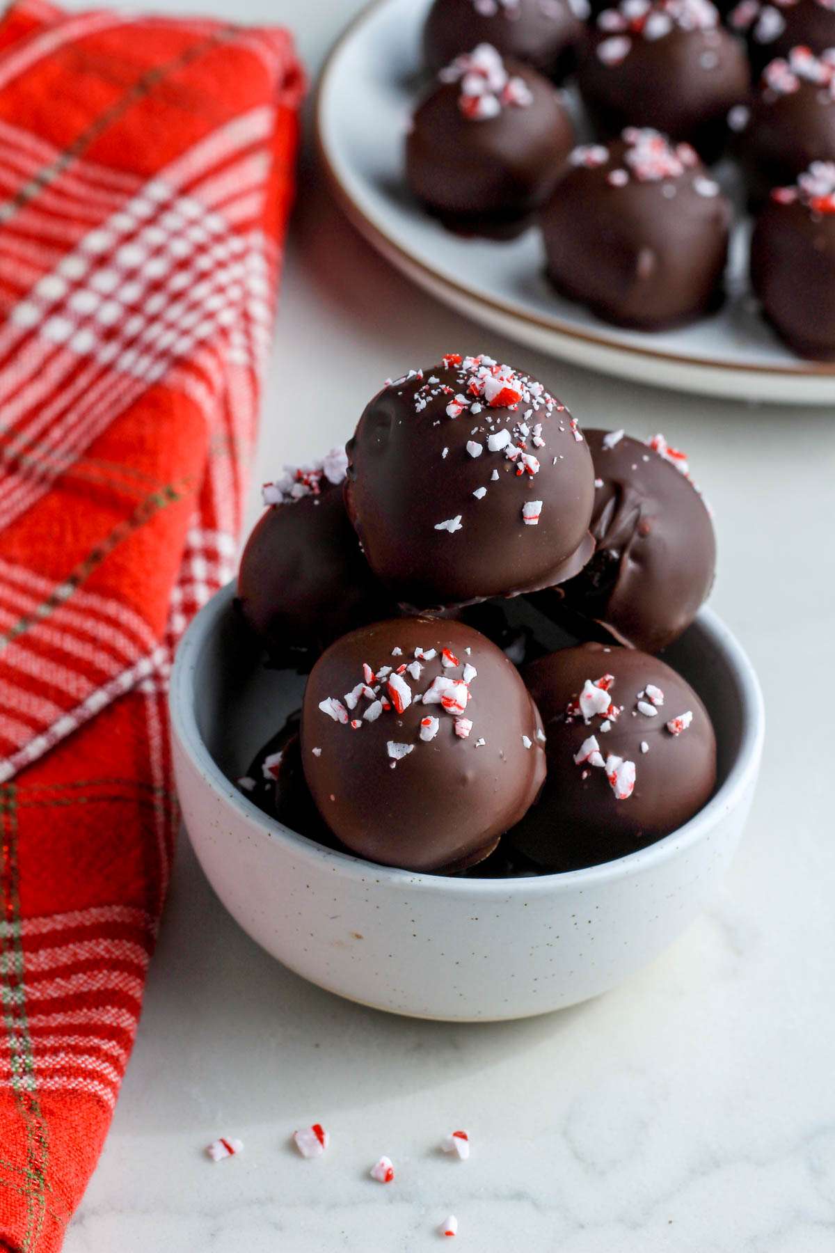 A small white bowl with peppermint Oreo truffles on a white counter with a plate of peppermint truffles in the back.