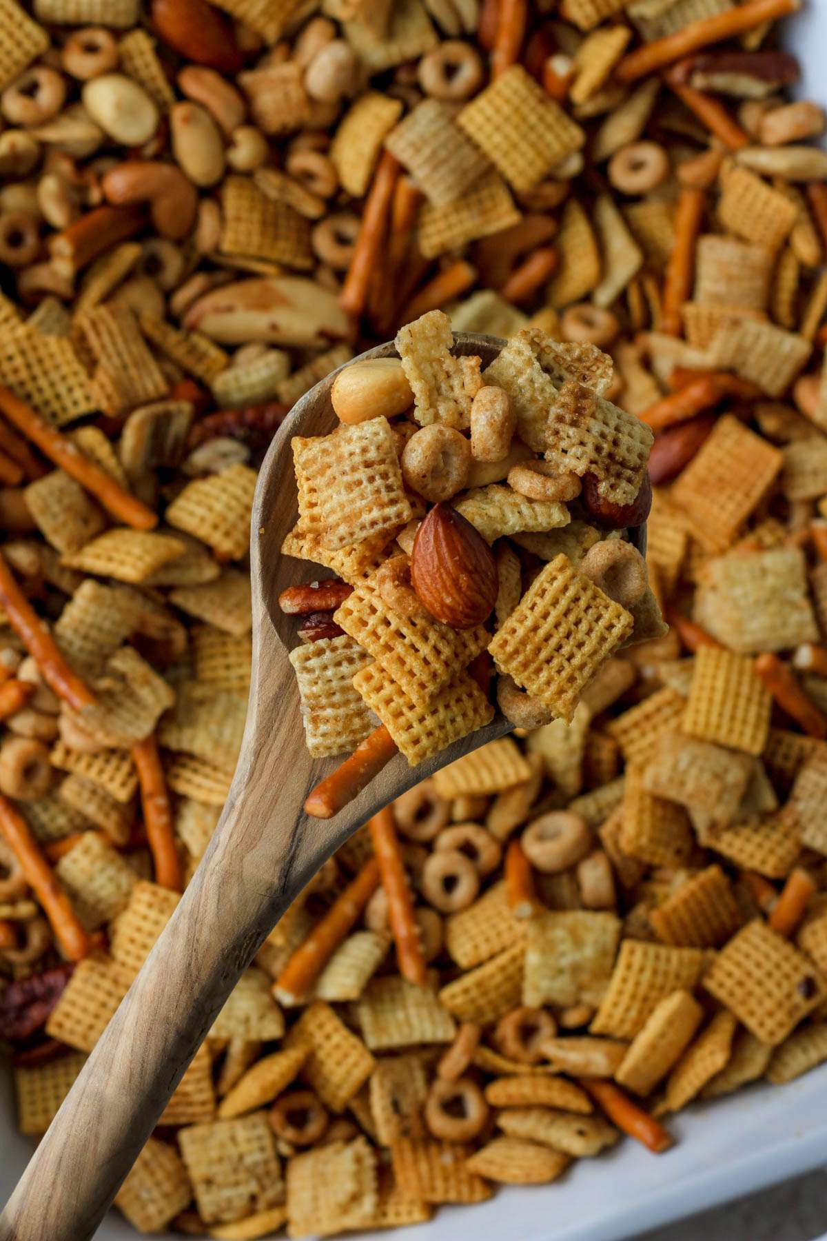 A wooden spoon holding a scoop of dairy-free Chex party mix over the baking dish filled with Chex party mix.