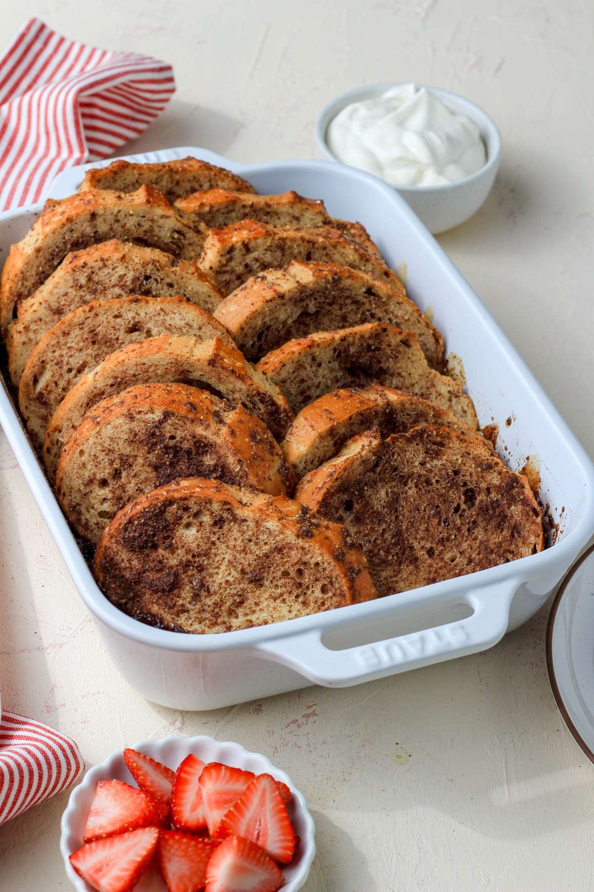 A white baking dish with vegan french toast bake and a bowl of strawberries and vegan whipped cream.