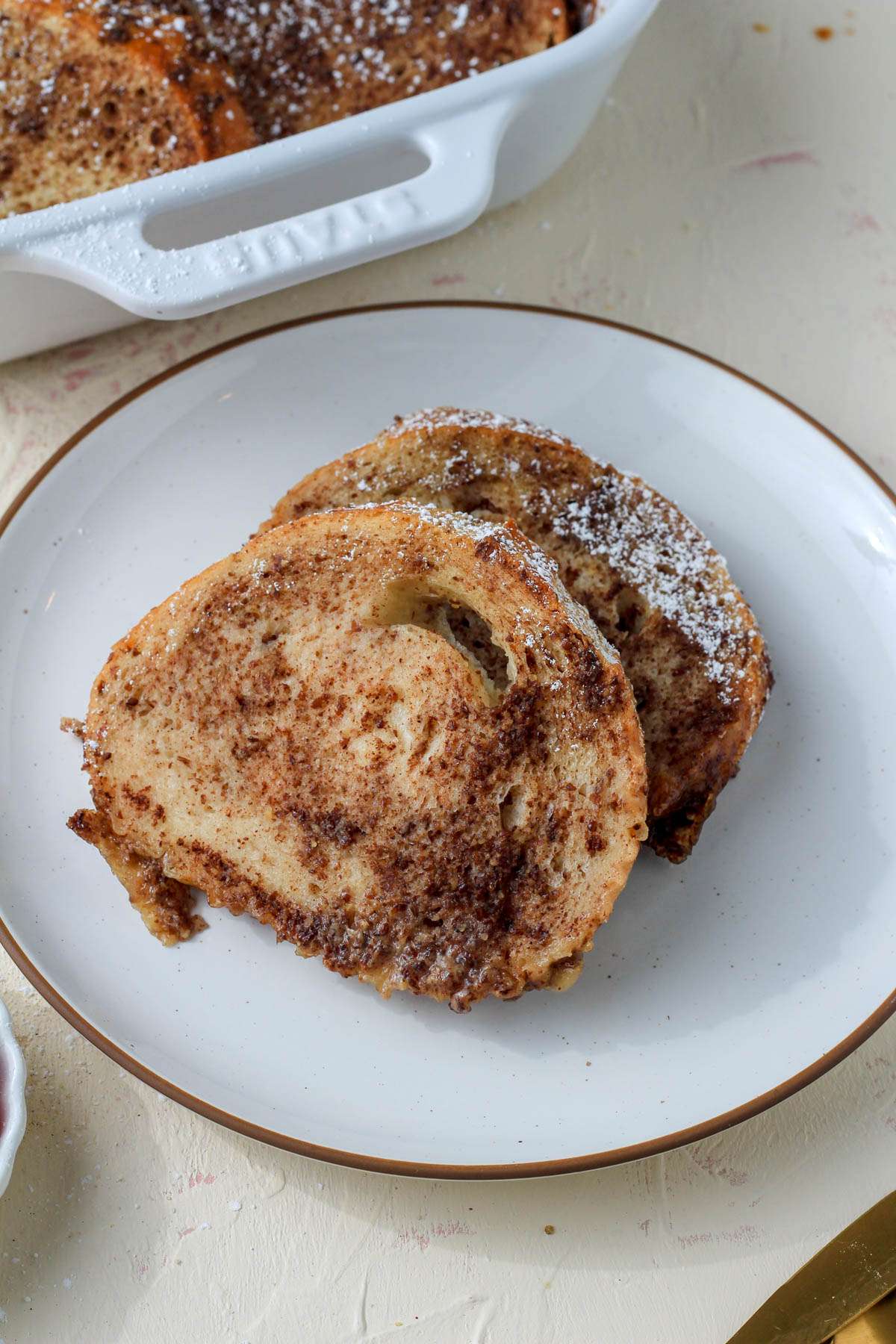 A white plate with a brown rim topped with two slices of vegan french toast and powdered sugar.
