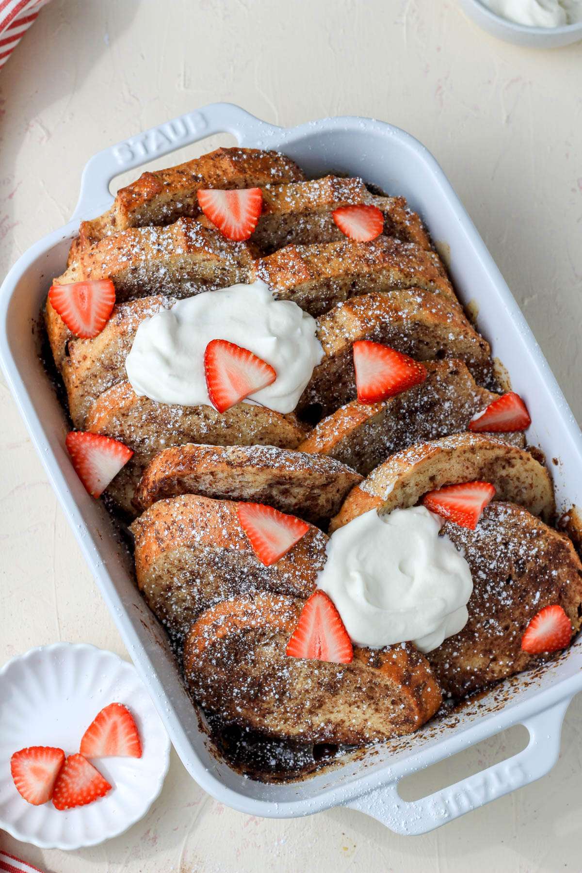 A white casserole pan with overnight vegan french toast bake topped with vegan whipped cream and strawberry slices.
