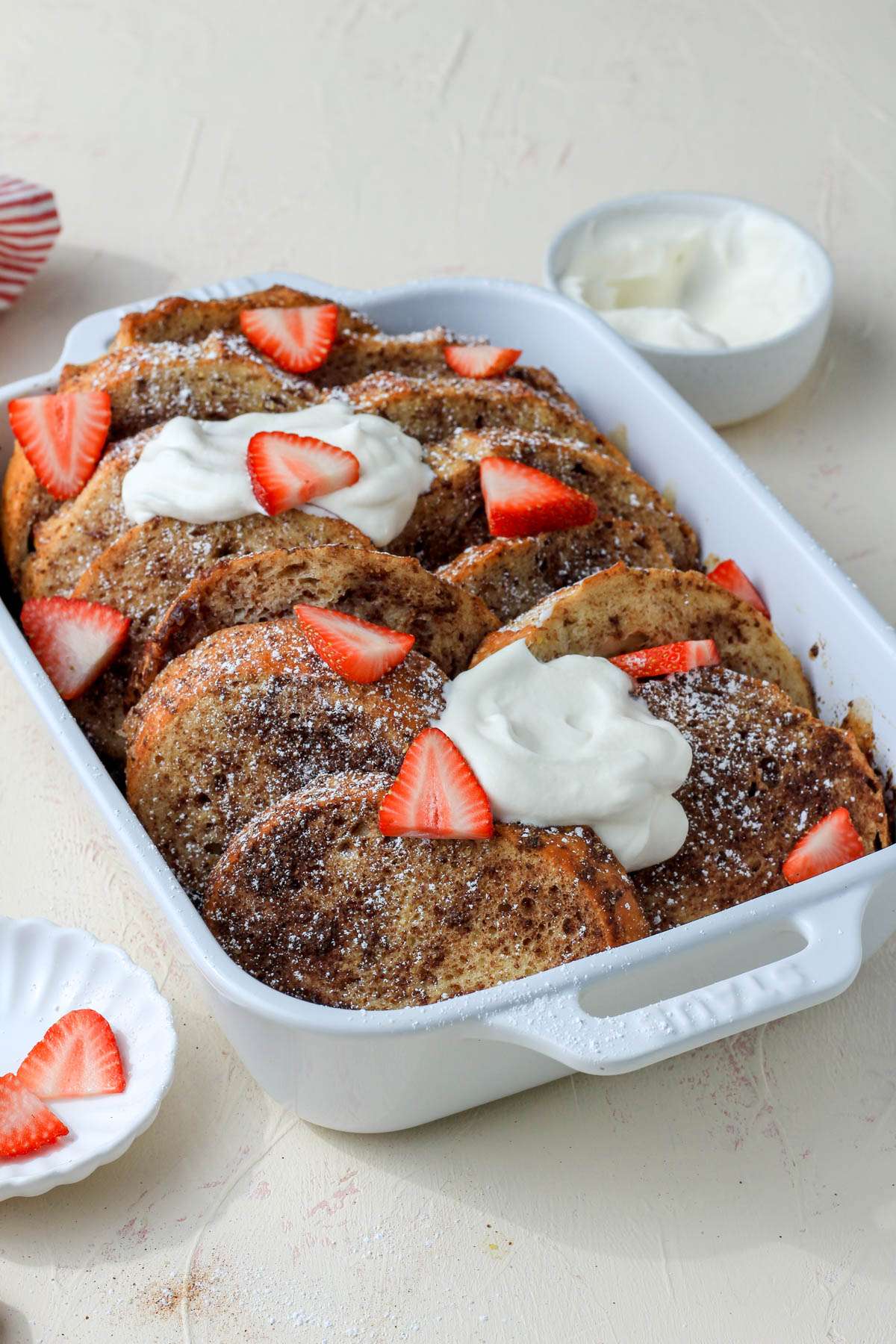 A white baking dish with overnight french toast topped with vegan whipped cream and sliced strawberries on a cream counter.