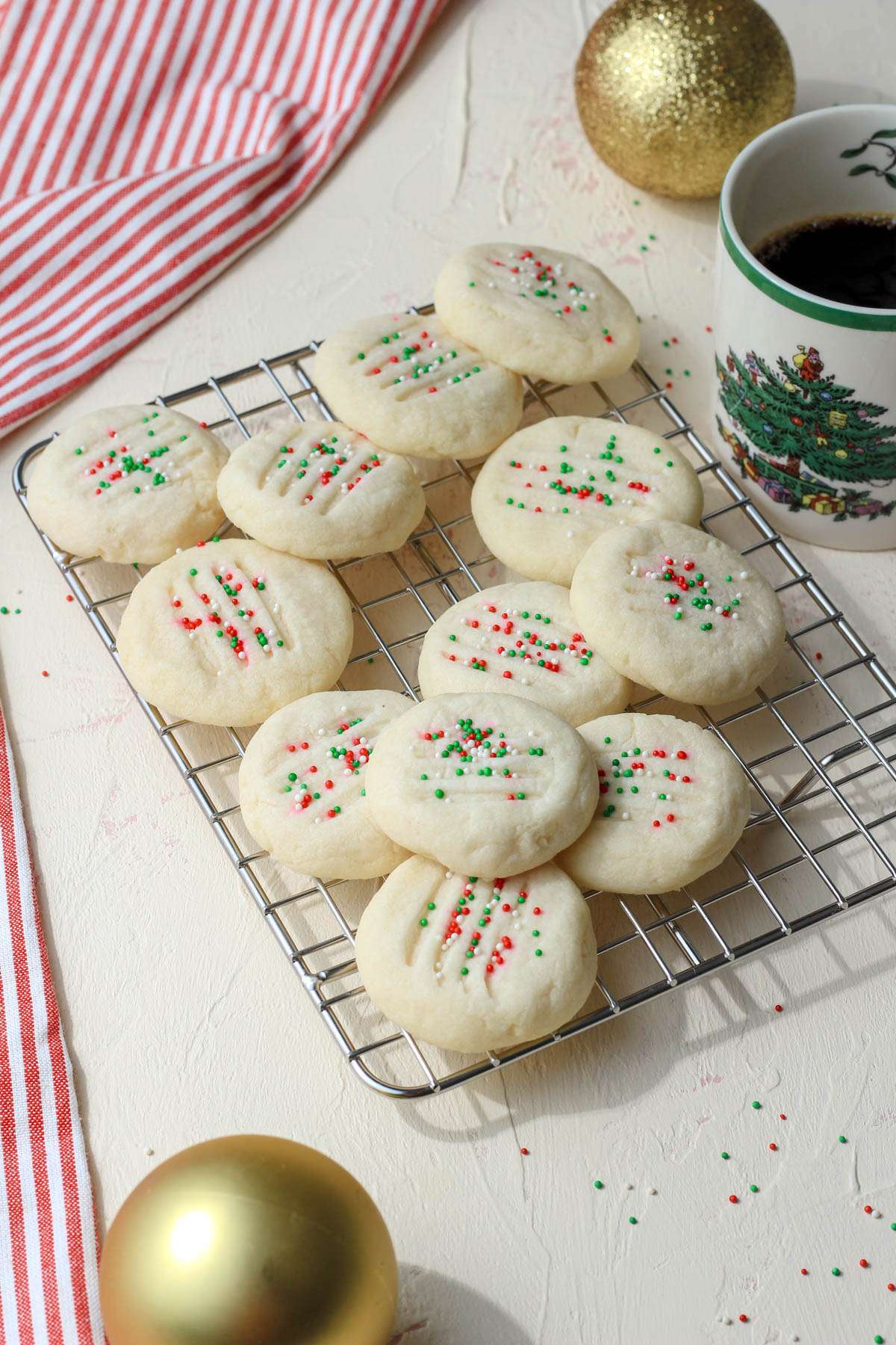 A wire rack with whipped shortbread cookies next to a cup of coffee and gold ornaments.
