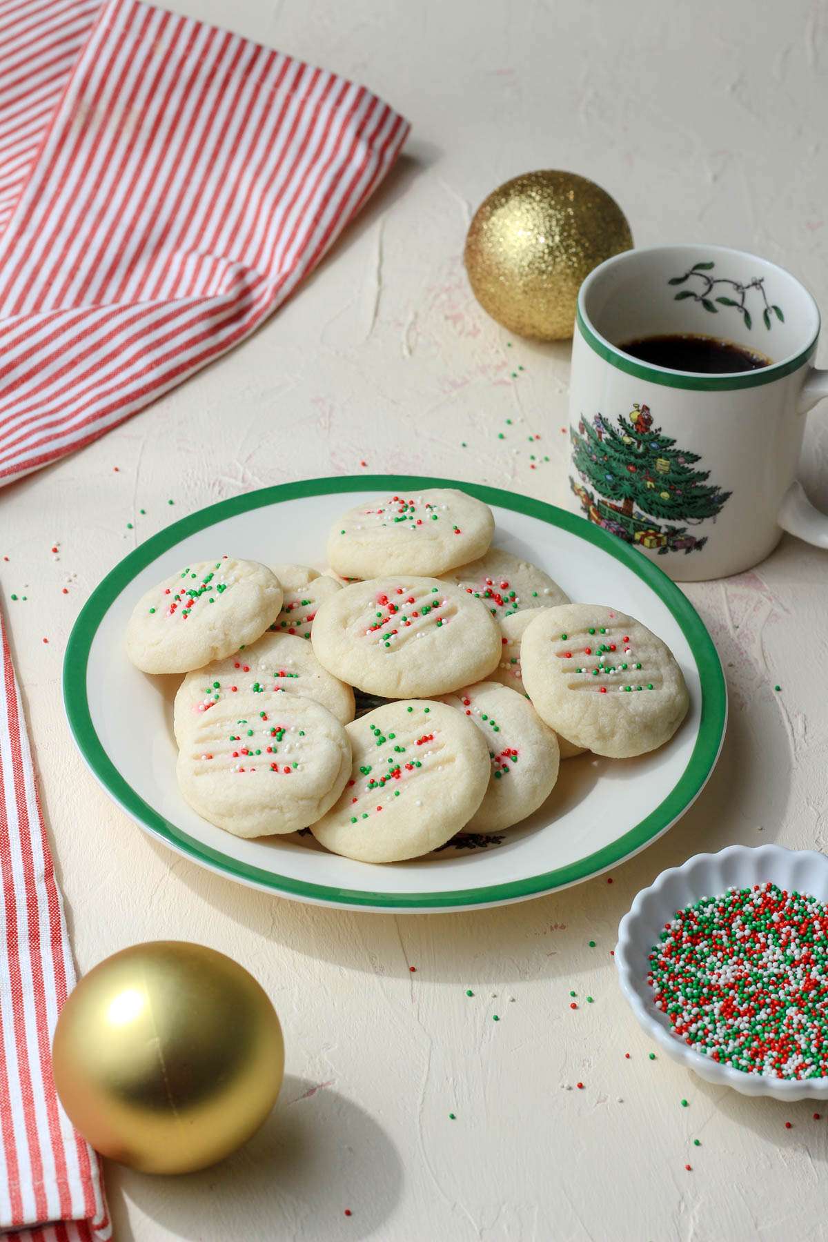 A white and green rimmed plate on a cream counter topped with vegan whipped shortbread cookies next to a mug of coffee.
