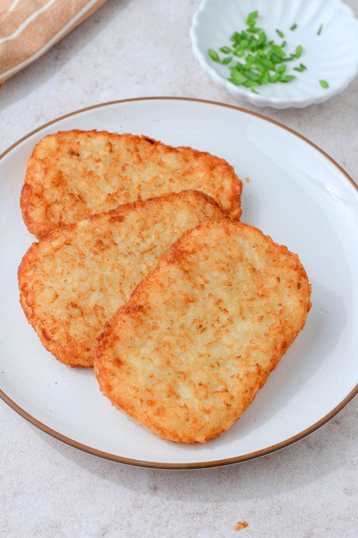 A white plate with a brown rim and three air fried hash browns next to a small white dish with cut green onions.