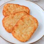 A close up of a white plate with a brown rim and three air fried frozen hash brown patties.