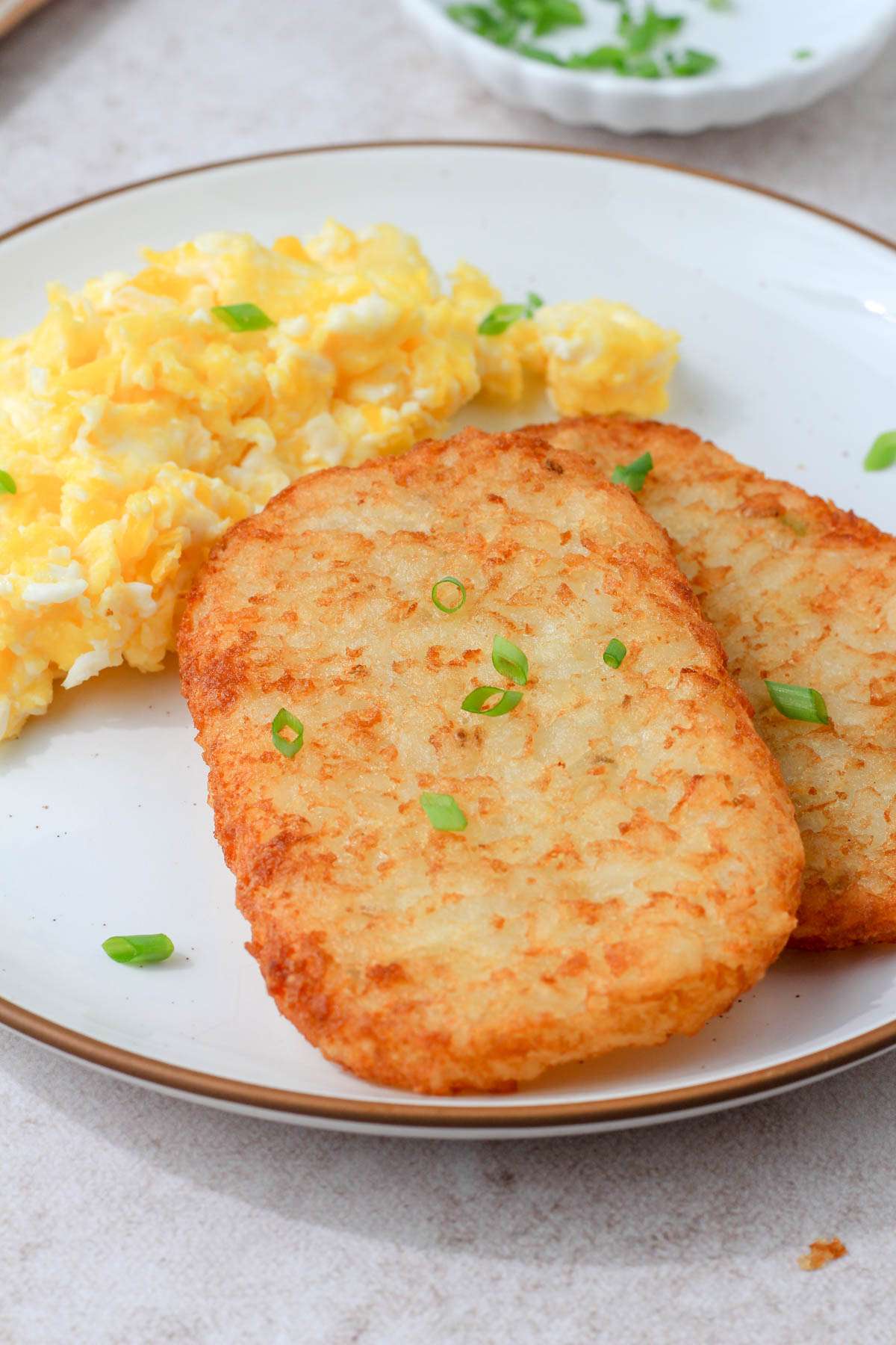 A top down image of a frozen hash brown patty topped with green onion in front of a small pile of scrambled eggs.