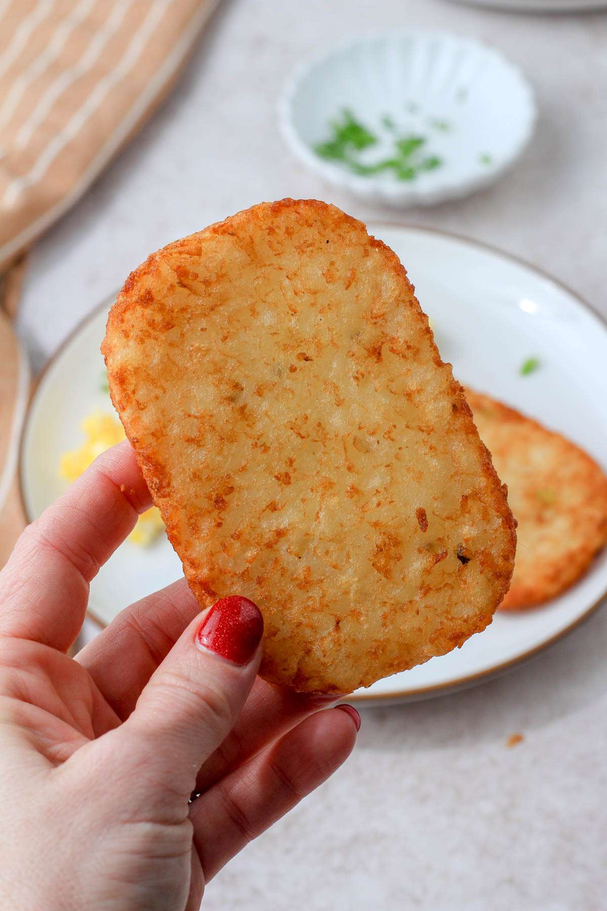 A hand holding an air fried hash brown patty in front of a white plate with a brown rim.