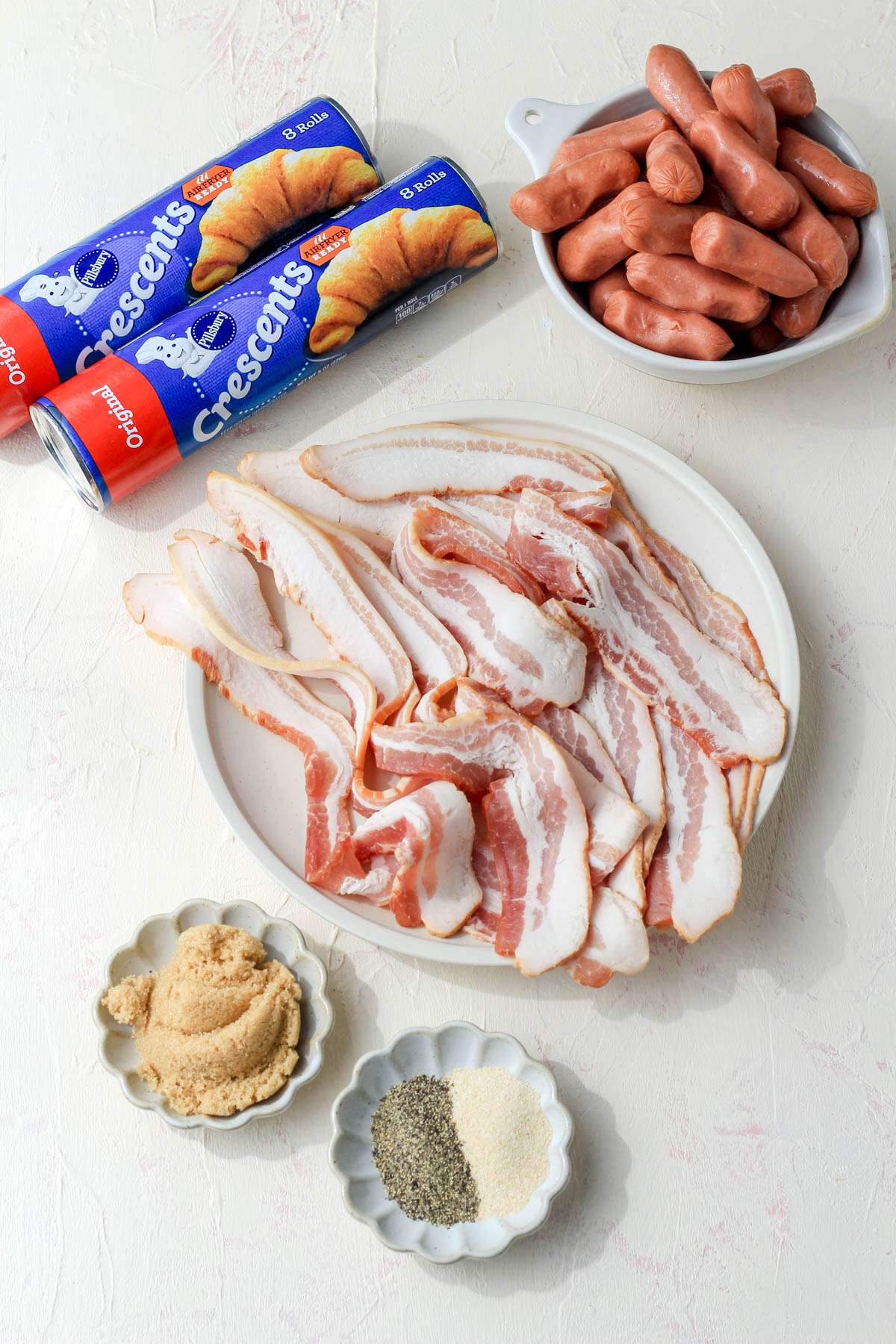 Ingredients for bacon wrapped pigs in a blanket on a cream and pink counter.