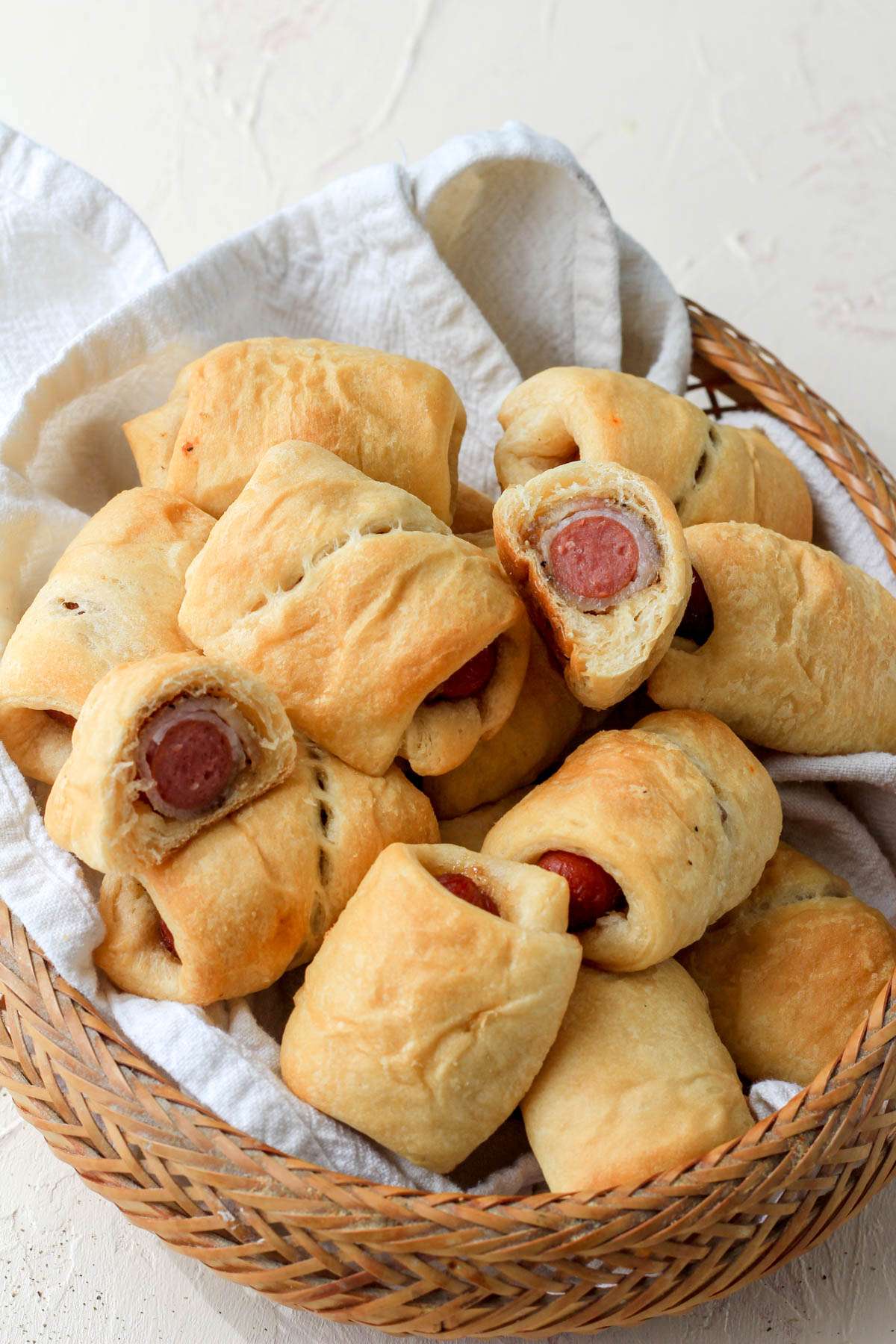 A brown basket with a white towel filled with bacon wrapped pigs in a blanket.
