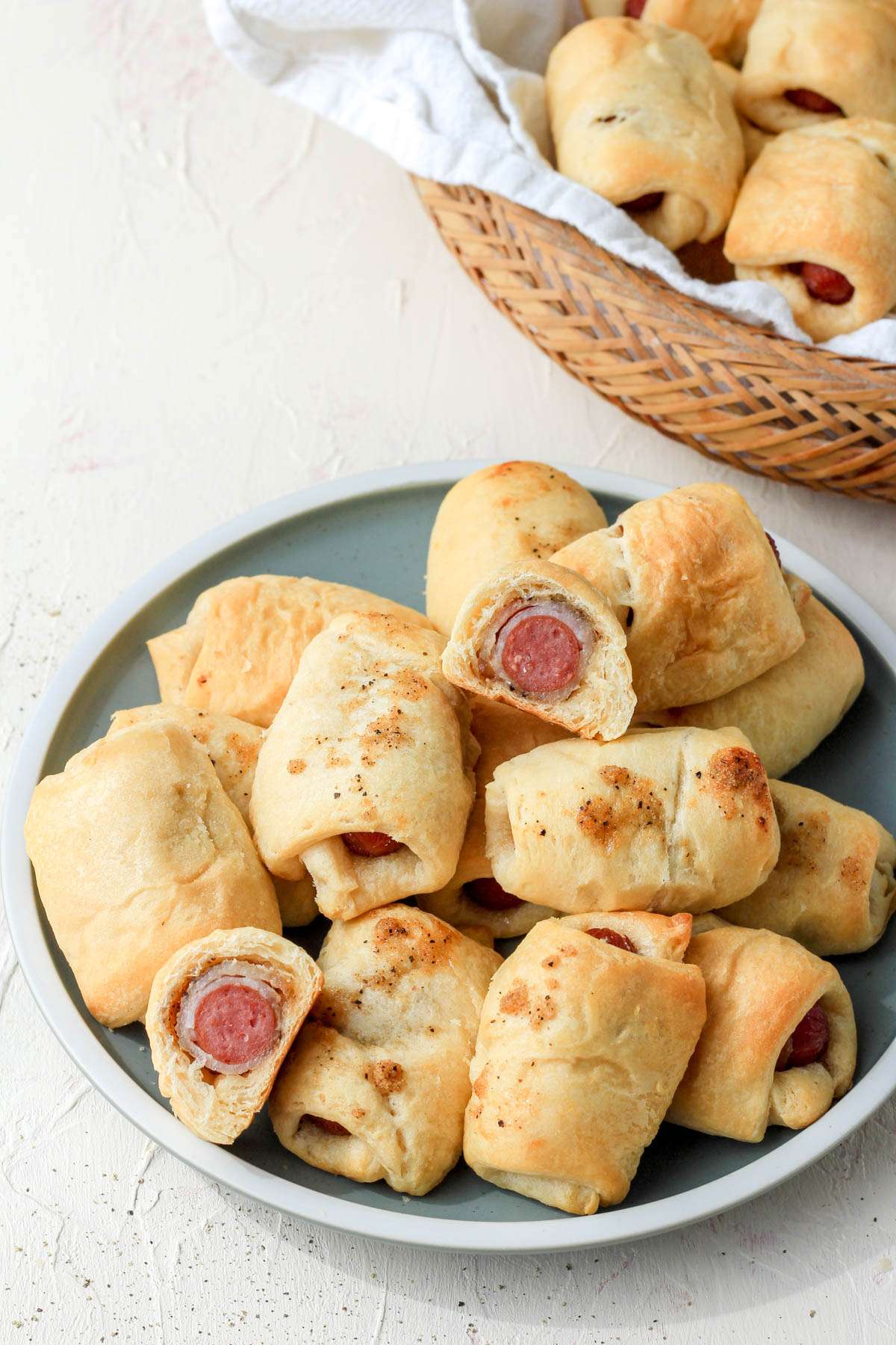 A green plate of bacon wrapped pigs in a blanket in front of a brown basket with bacon pigs in a blanket.