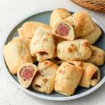 A green plate with a white rim topped with various bacon wrapped pigs in a blanket with one sliced in half to show the inside.