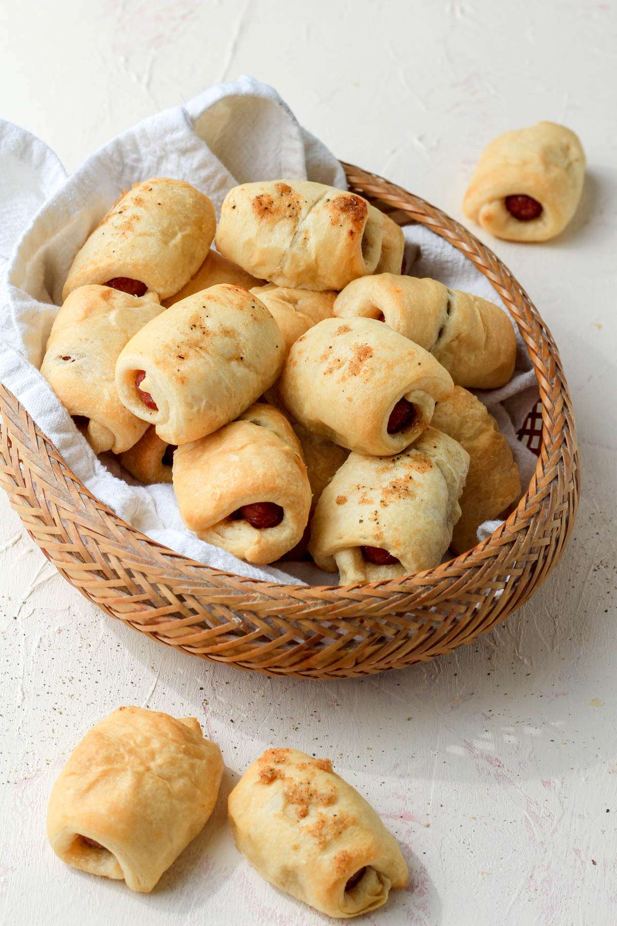 A brown basket with a white dish towel filled with bacon wrapped pigs in a blanket with a few on the cream counter in front.