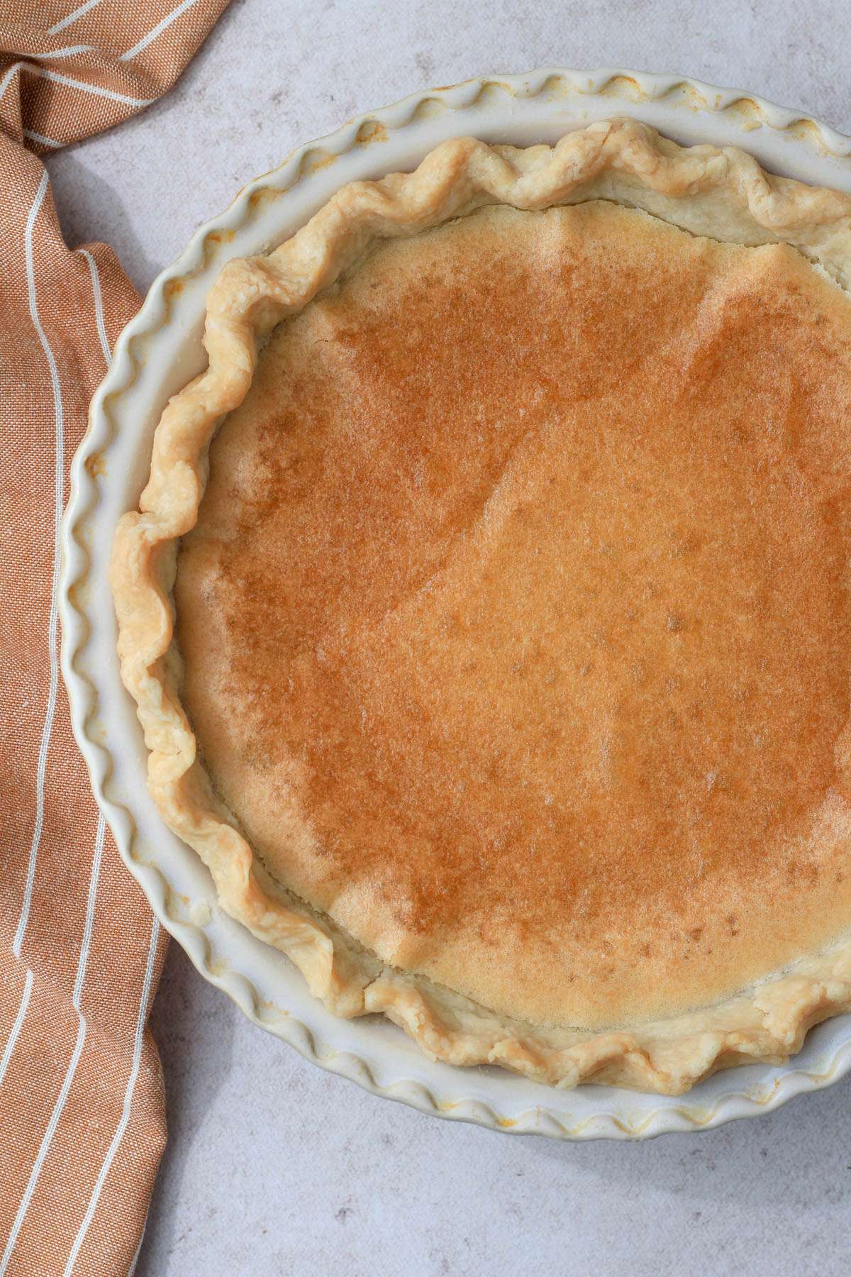 A close up of a chess pie in a cream pie pan with a rust colored dish towel to the left.