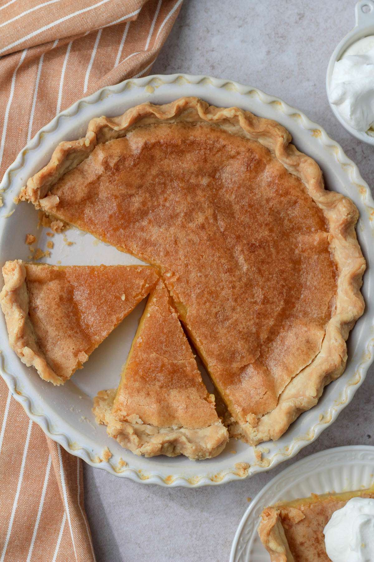 A cream pie pan with dairy-free chess pie sliced into wedges with a small bowl of whipped cream in the top right and a small plate with a slice of pie in the bottom right.