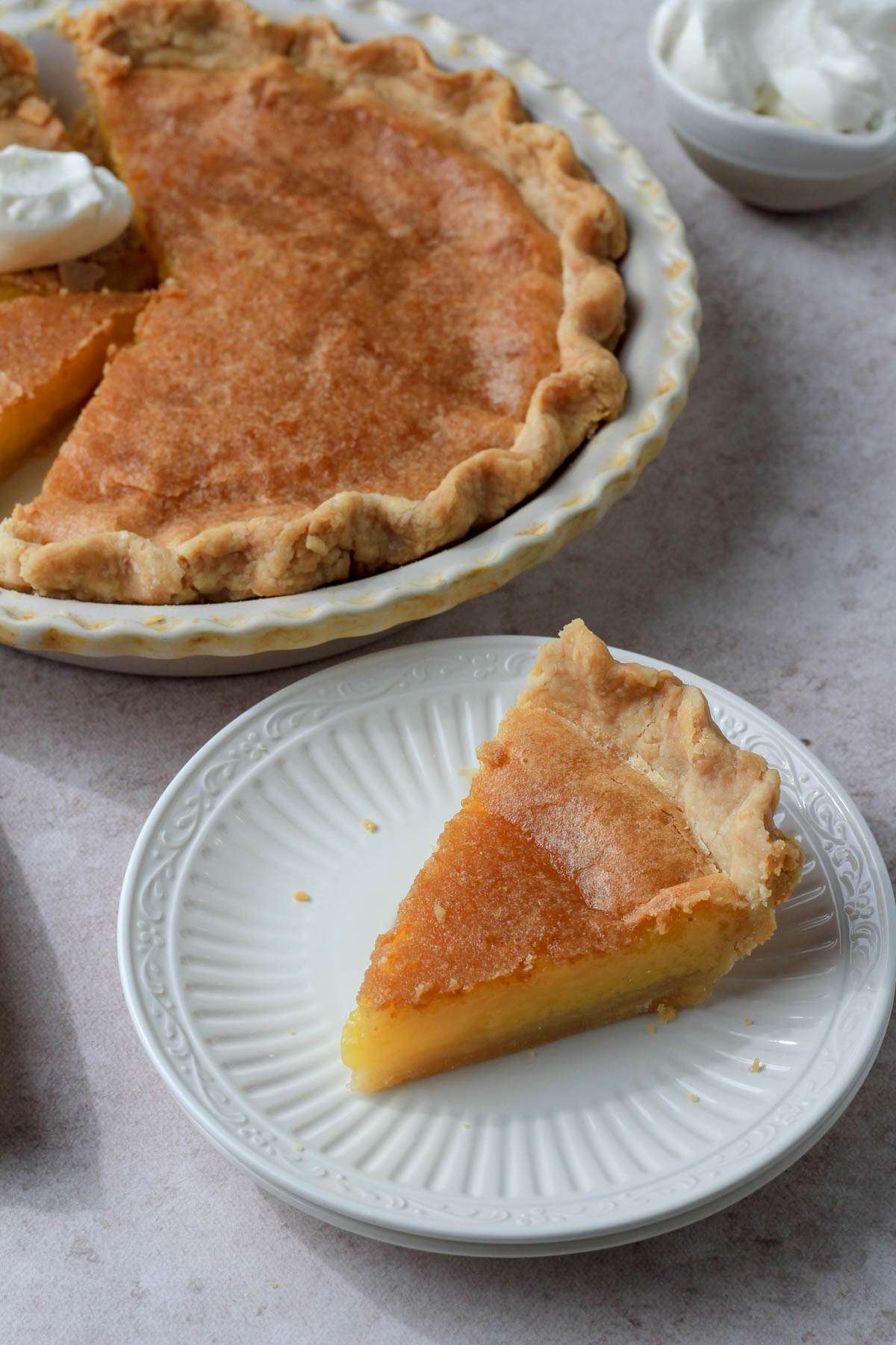 A cream plate with a slice of dairy-free chess pie in front of a cream pie pan with chess pie and a small bowl of vegan whipped cream in the back right corner.