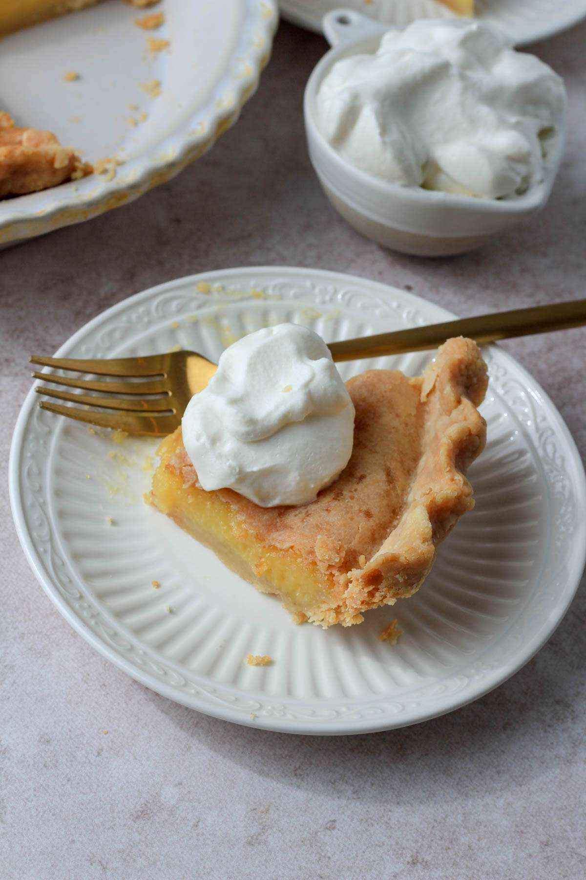 A slice of chess pie topped with vegan whipped topping on a small plate with a bite missing and a gold fork behind the slice.
