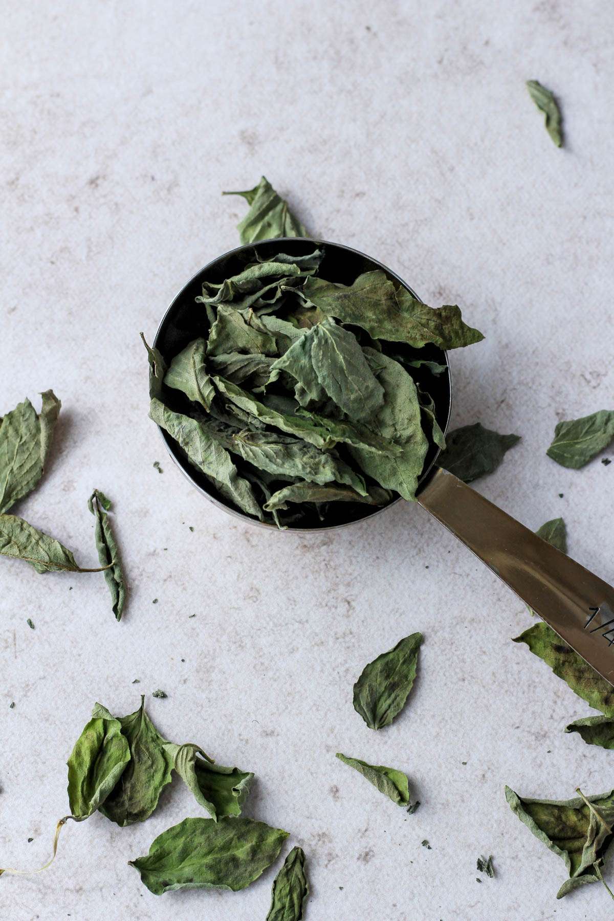 A gold measuring cup with dried basil leaves before crushing.