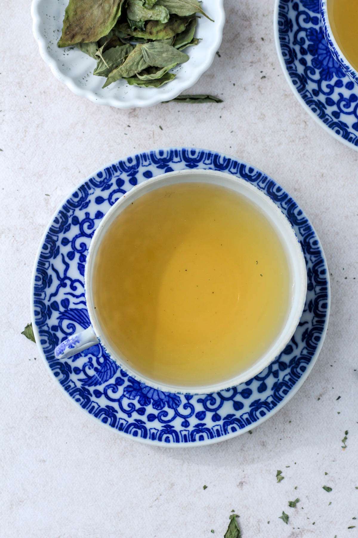A top down image of dried basil tea in a blue and white cup and saucer on a tan counter.