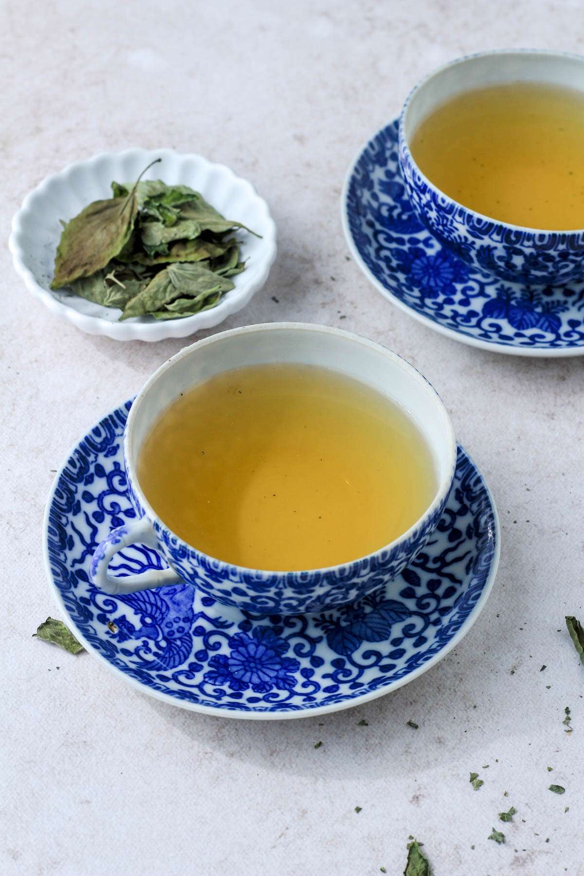 Two cups of dried basil tea on a tan counter with a small white dish of dried basil in the back left.
