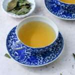 A cup and saucer of dried basil tea in front of a small white plate of dried basil.