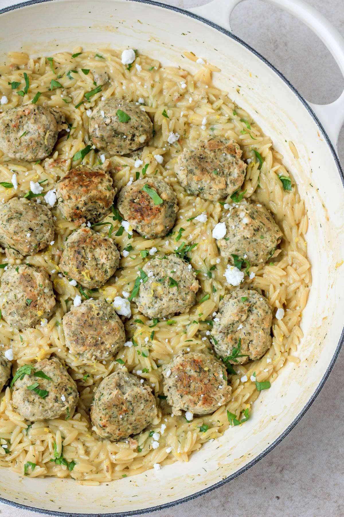 The right side of a white skillet with Greek turkey meatballs with creamy lemon orzo on a cream counter.