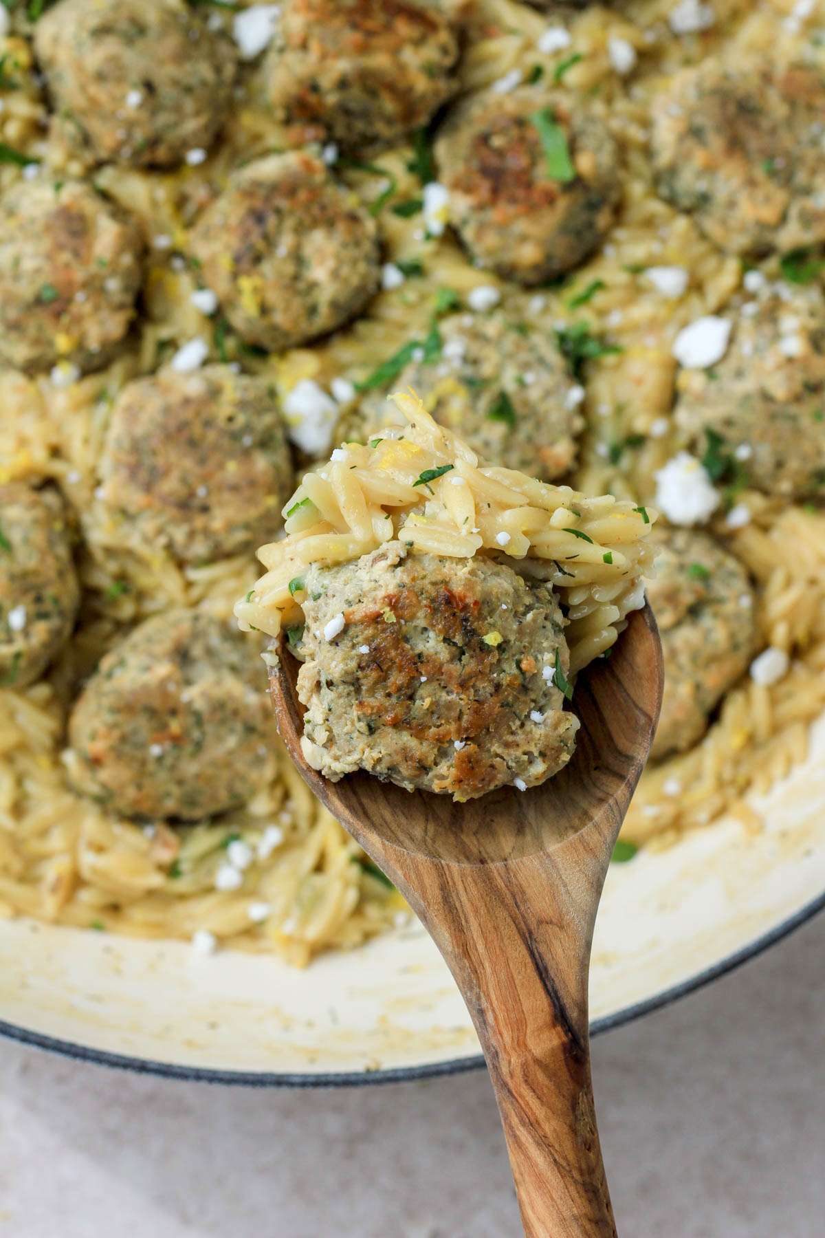 A wooden spoon with a scoop of creamy lemon orzo and a greek turkey meatball hovering over a white skillet.