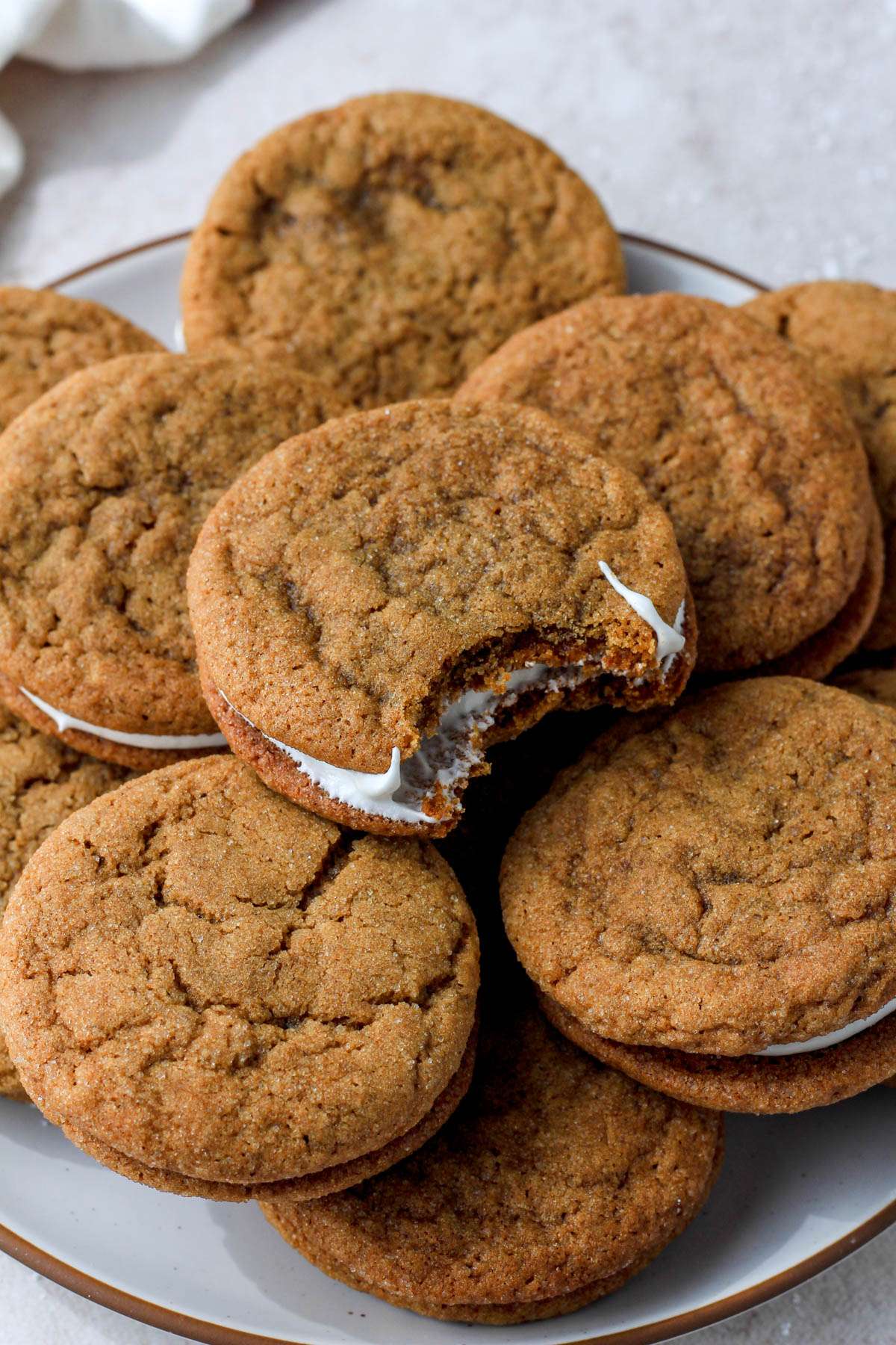 A white plate with a brown rim and a pile of soft ginger cookie sandwiches with the top cookie sandwich having a bite out.