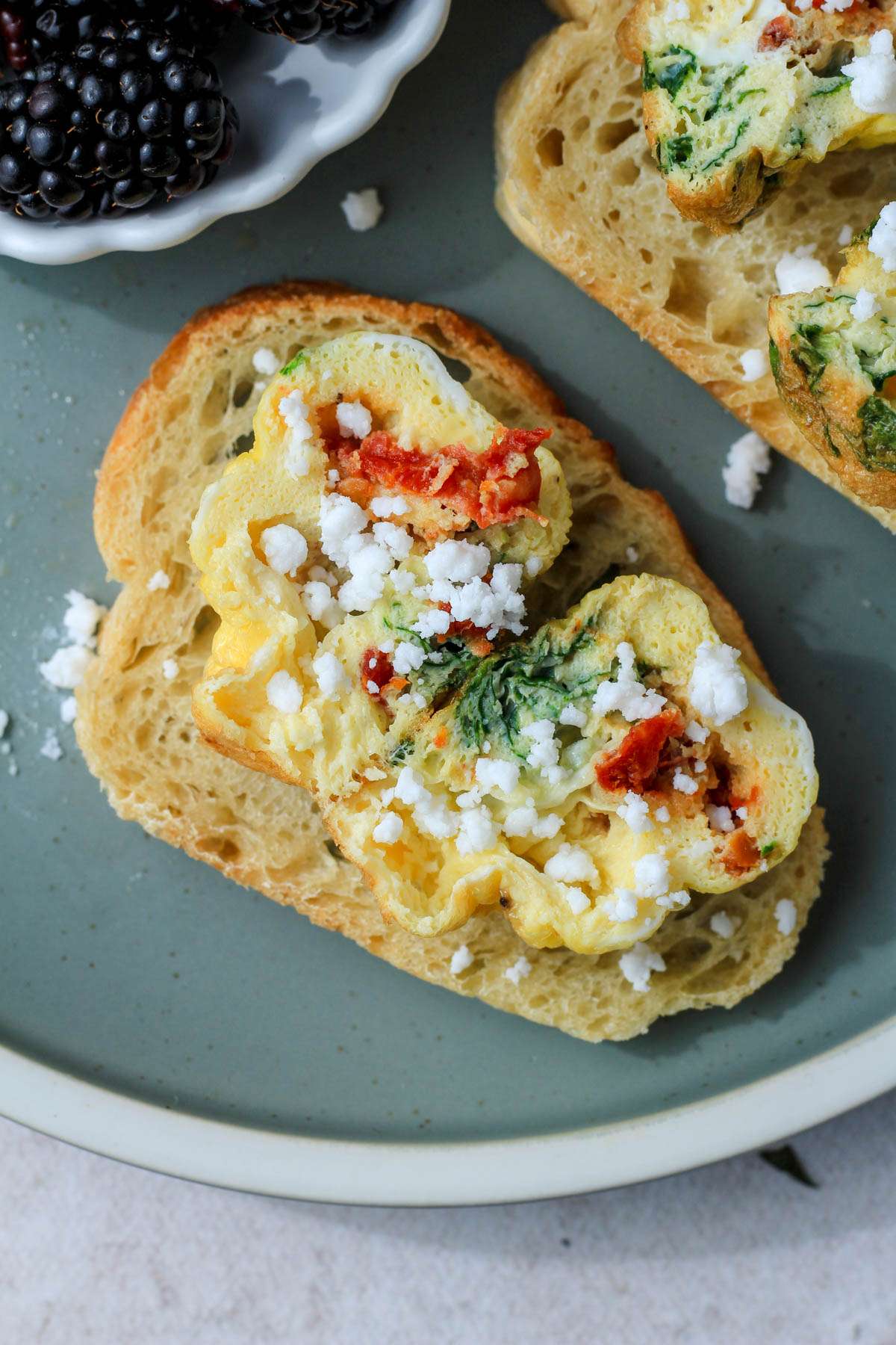 A green plate with a white rim and a slice of bread topped with a cut open spinach, feta, and sun dried tomato egg bite sprinkled with dairy-free feta.