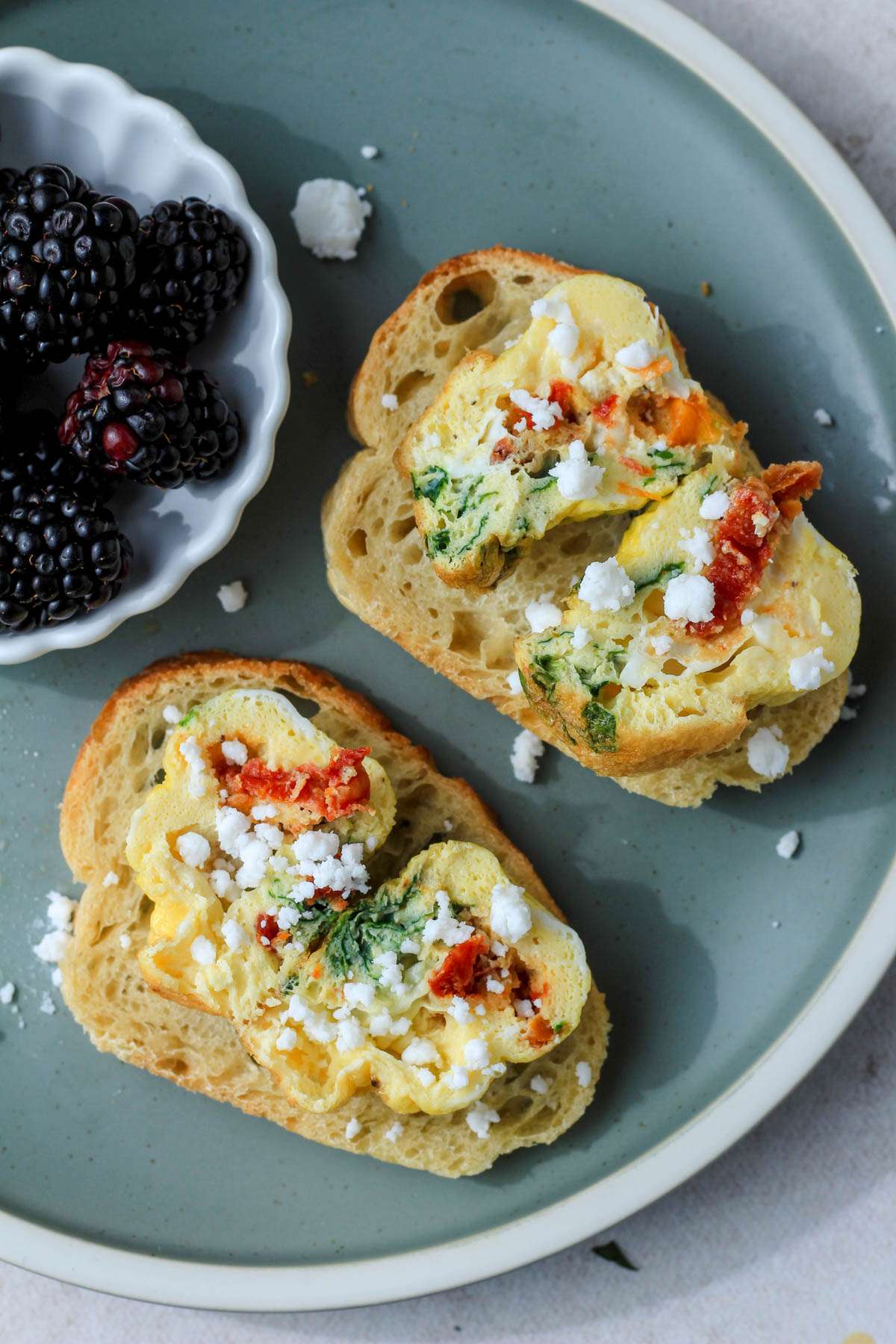 Two slices of bread on a green plate with spinach, feta, and sun dried tomato egg bites sprinkled with dairy-free feta next to a small plate of blackberries.