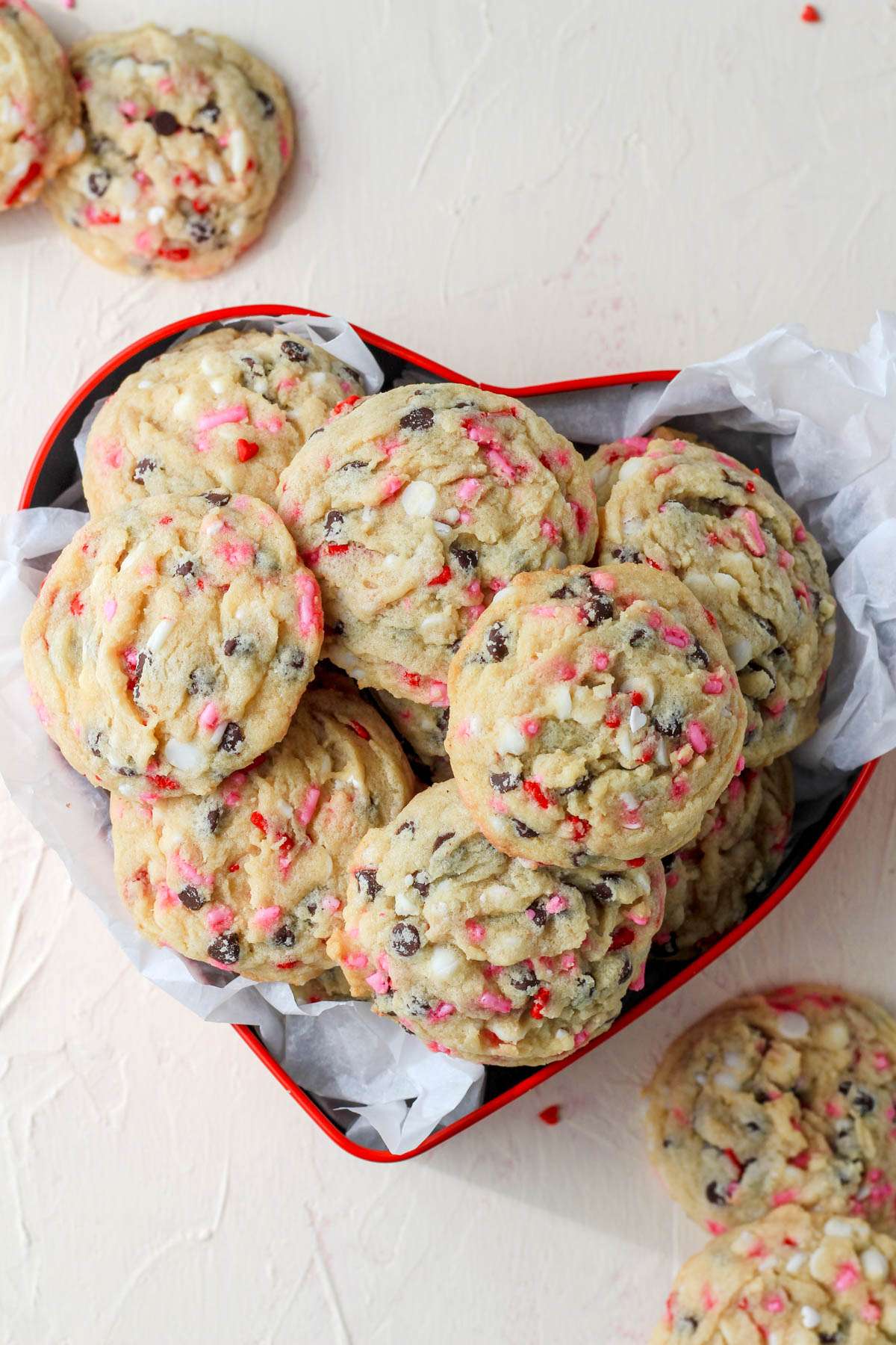 A red cookie tin with parchment paper and a pile of valentines day chocolate chip cookies on a cream counter.