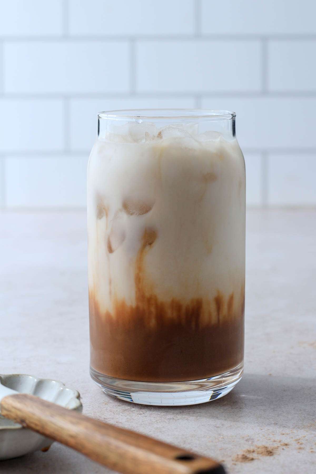 A chocolate almond milk shaken espresso with protein powder in a tan counter with a wooden handled spatula in the front.