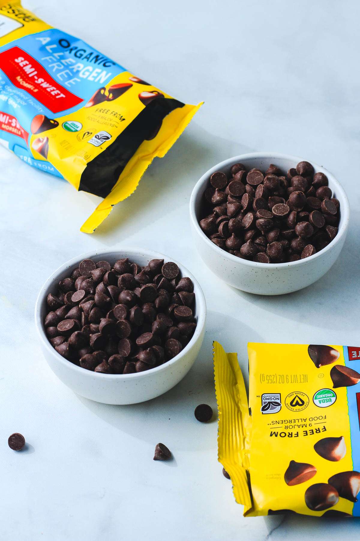 Two white bowls of nestle toll house dairy-free chocolate chips next to open bags of chocolate chips.