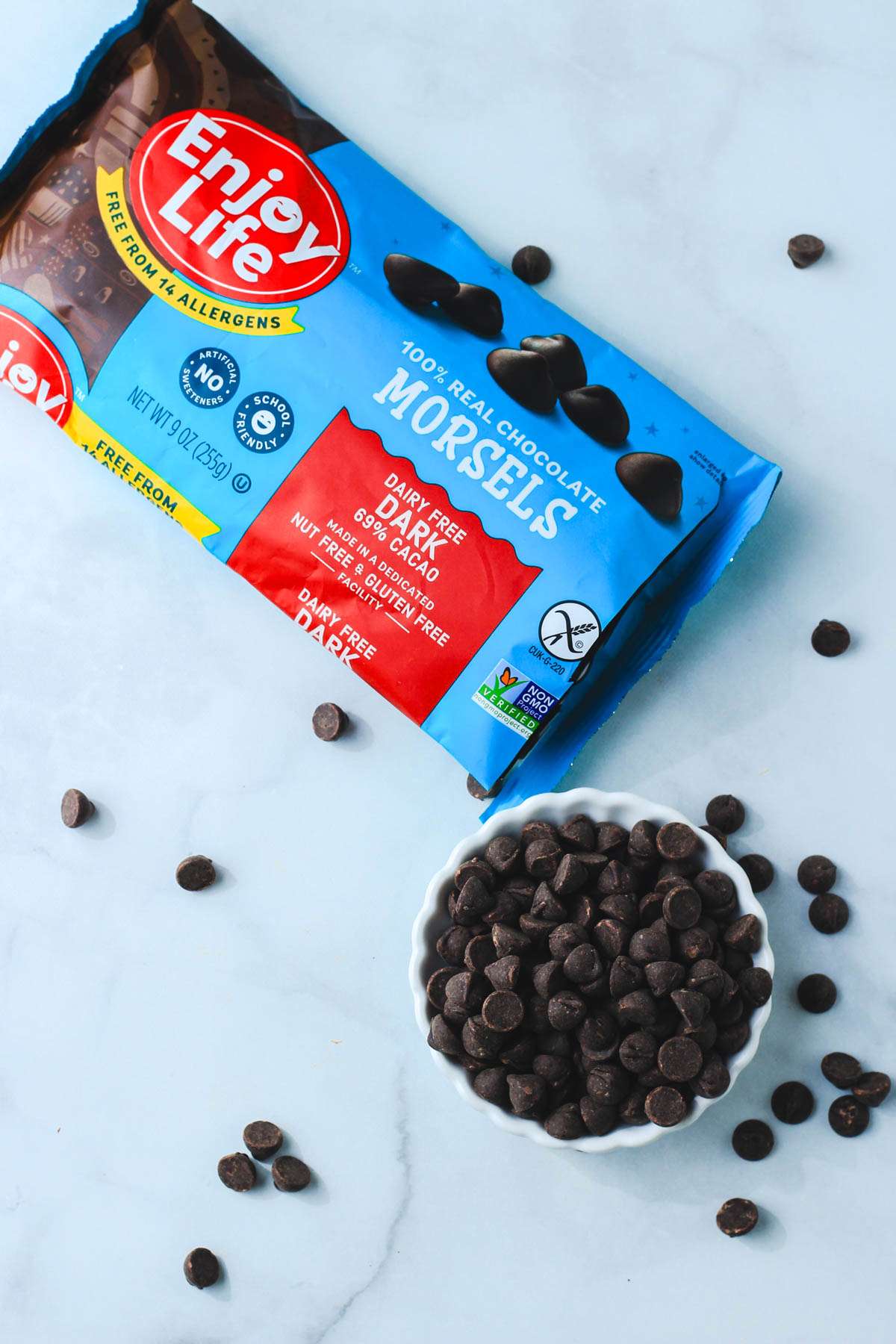A blue bag of enjoy life dark chocolate morsels poured into a small white bowl on a grey counter.