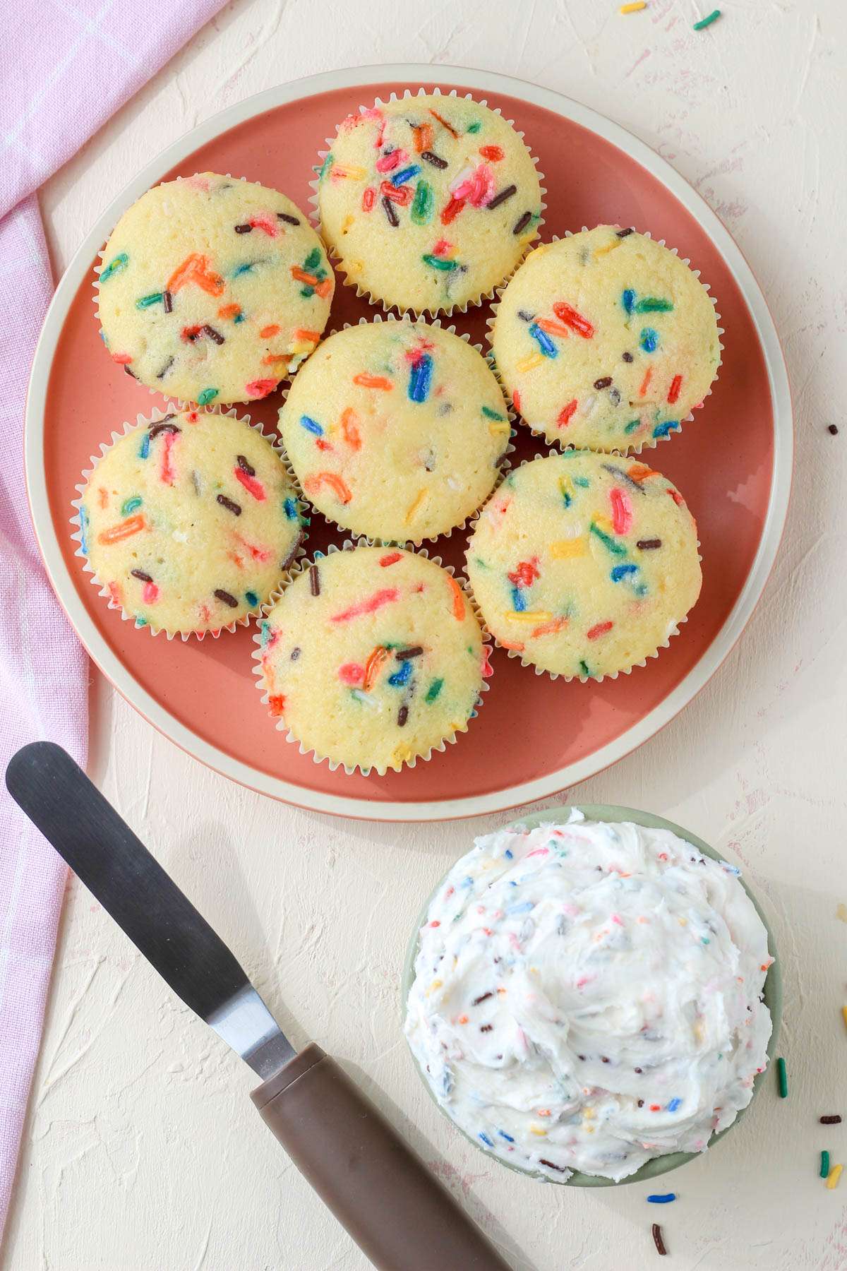 A round pink plate in the top left with a small green bowl of dairy-free funfetti icing in the bottom right.