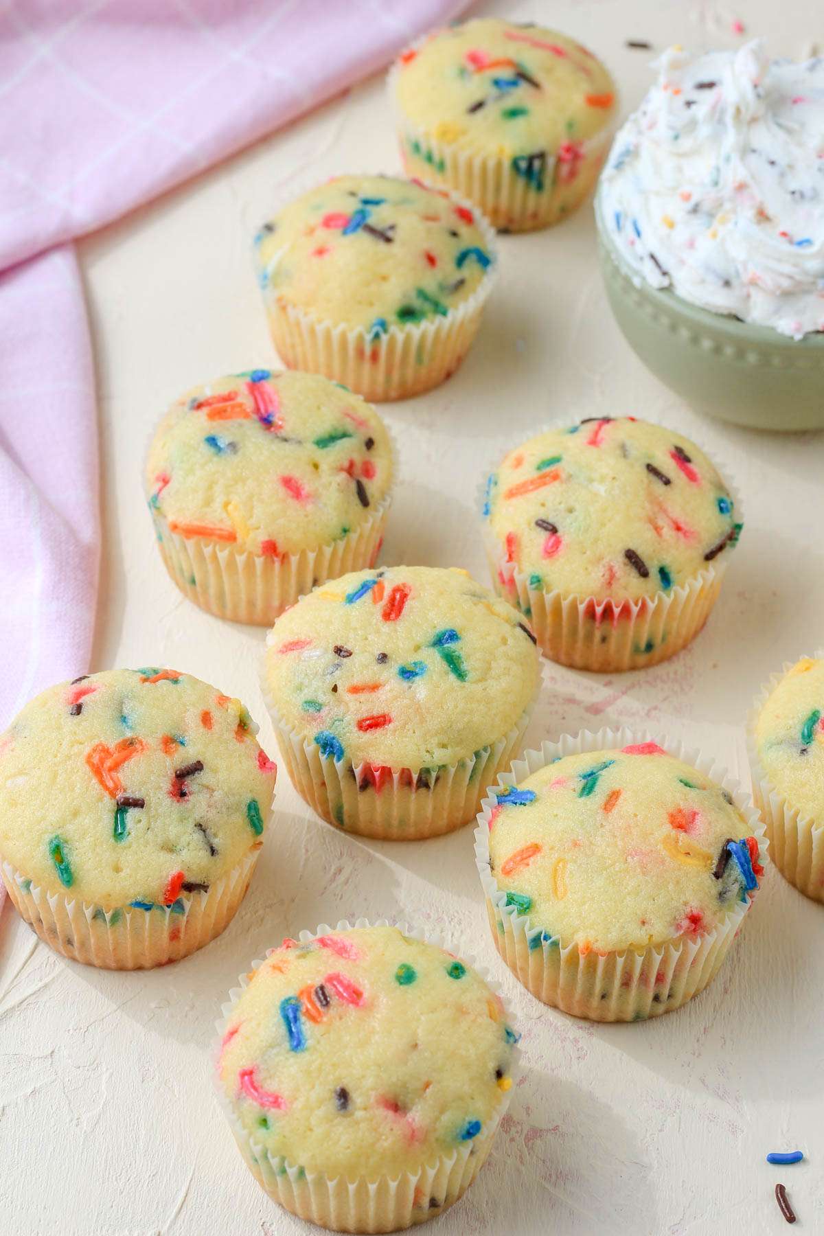 A cream and pink counter with dairy-free funfetti cupcakes scattered on the counter with a small green and white bowl of icing in the back right corner.