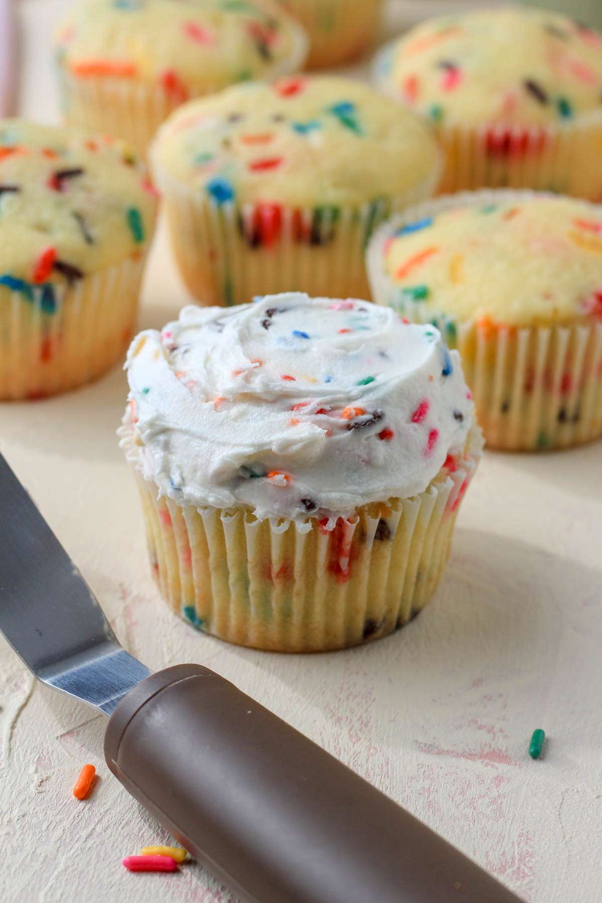 An offset spatula in front of a dairy-free funfetti cupcake topped with dairy-free buttercream icing.